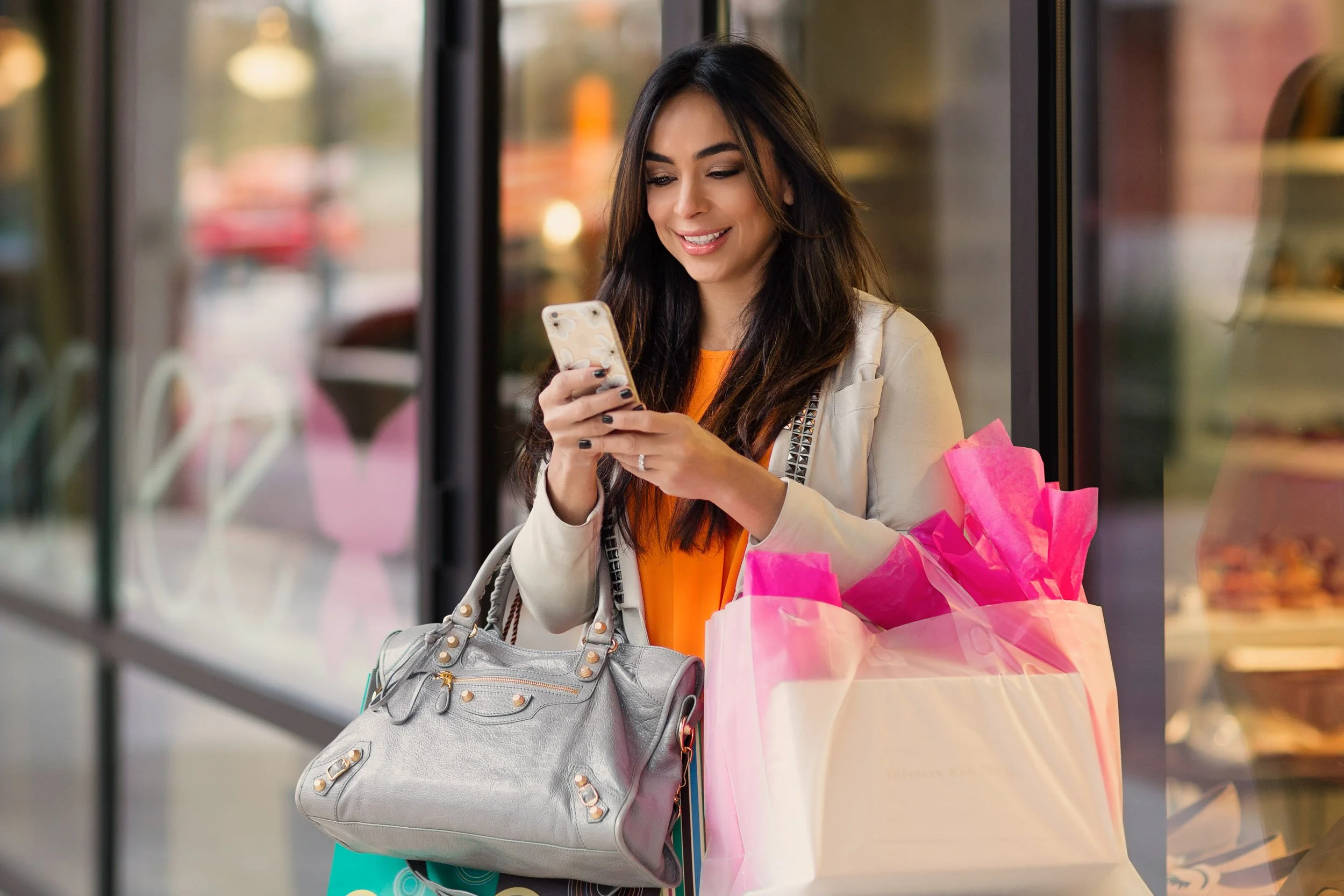 A woman with long dark hair, wearing an orange top and beige jacket, holding a smartphone, standing next to shopping bags and a gray purse, inside a store or shopping mall.
