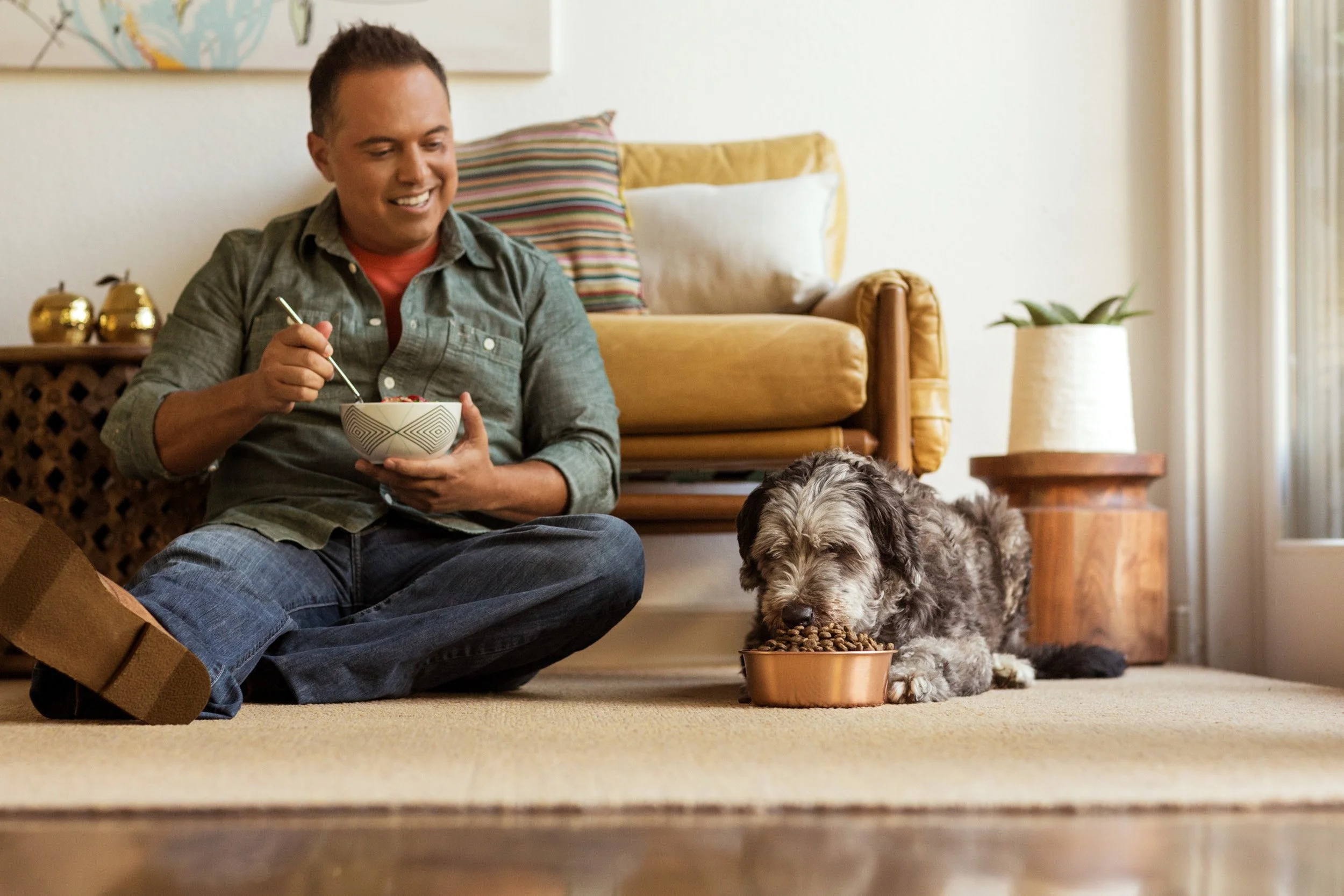 A man sitting on the floor, smiling, holding a bowl of cereal or snack, with a dog lying nearby eating from a bowl, in a cozy living room with a yellow couch, striped pillows, potted plant, and a rug.