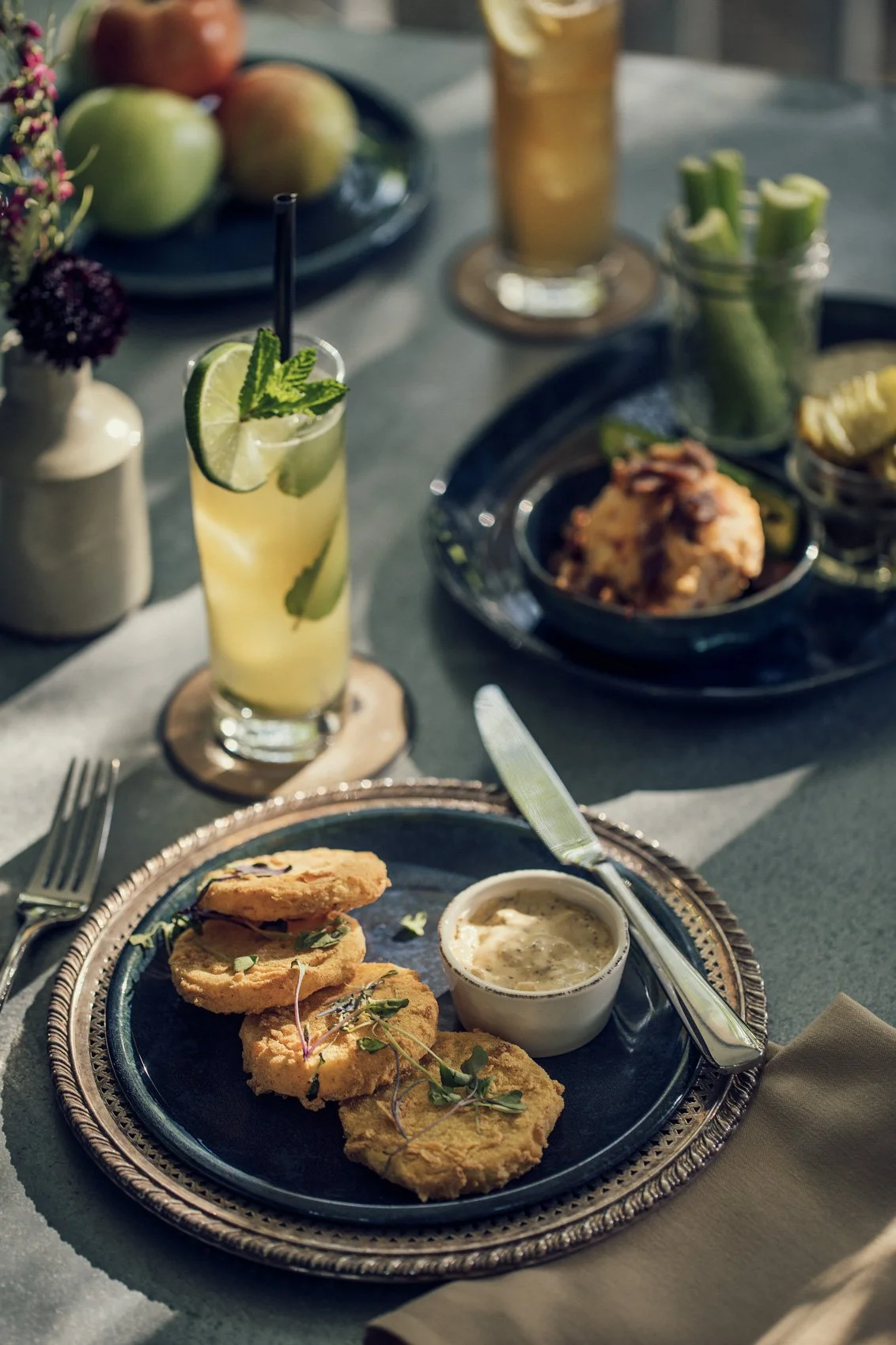 Fried fish fillets served with tartar sauce on a decorative plate, a glass of lemonade garnished with lime and mint, and various side dishes including celery sticks, a bowl of fried chicken, crackers, and apples in the background on a dining table.