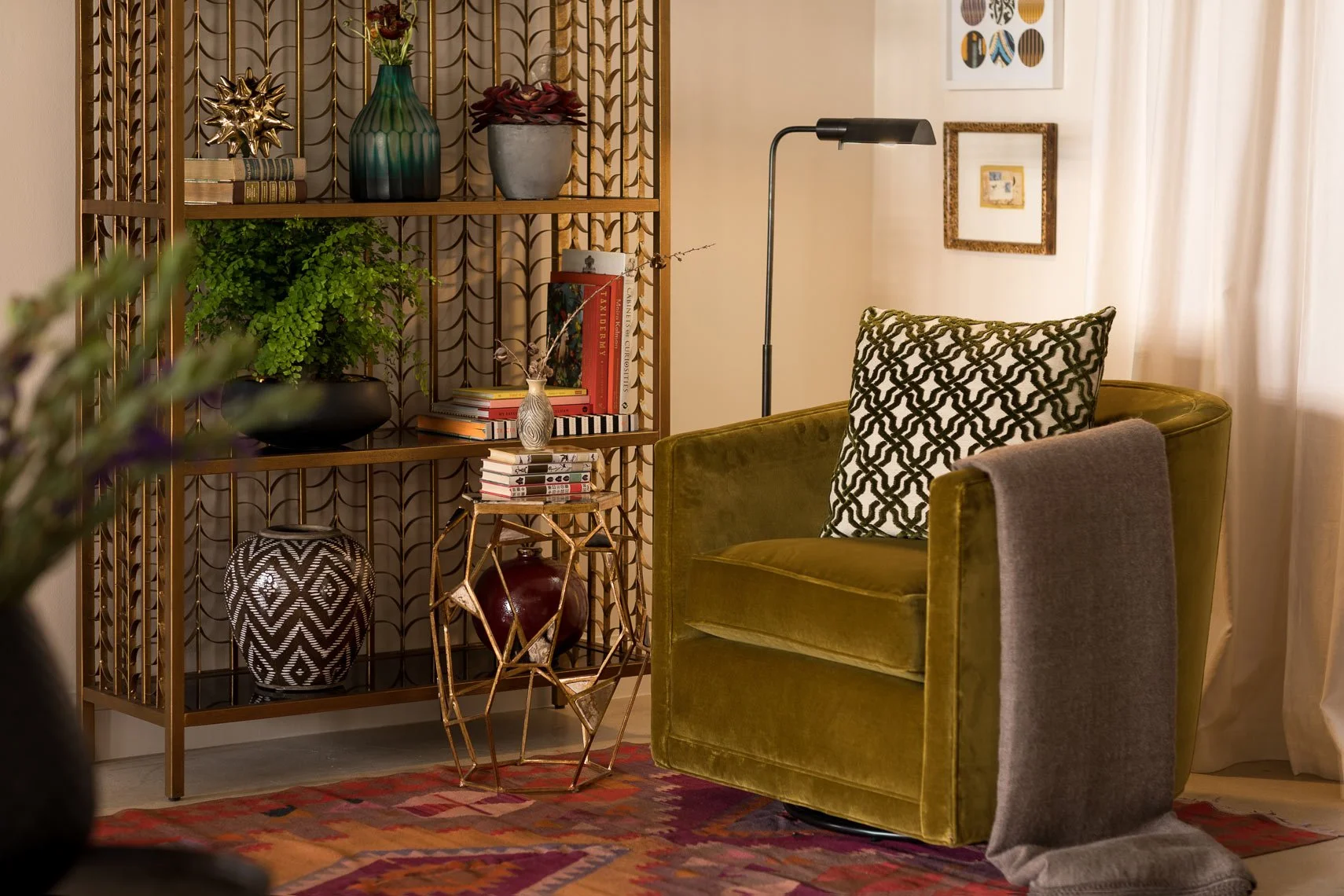 Living room with an olive green velvet armchair with a black and white patterned pillow and a gray blanket draped over the armrest. Wooden shelves with decorative objects including vases, books, and plants. A small side table with a decorative geomet