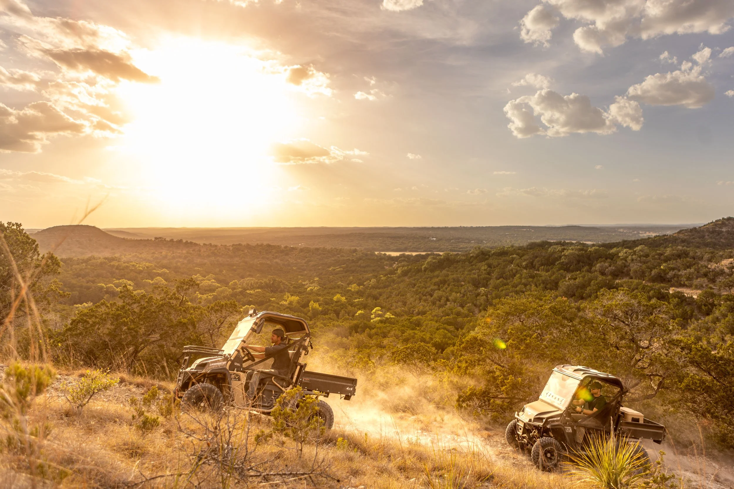 Two people riding in off-road utility vehicles on a dusty trail through a hilly, wooded landscape during sunset or sunrise. The sky is partly cloudy with the sun low on the horizon, creating a warm, golden glow.