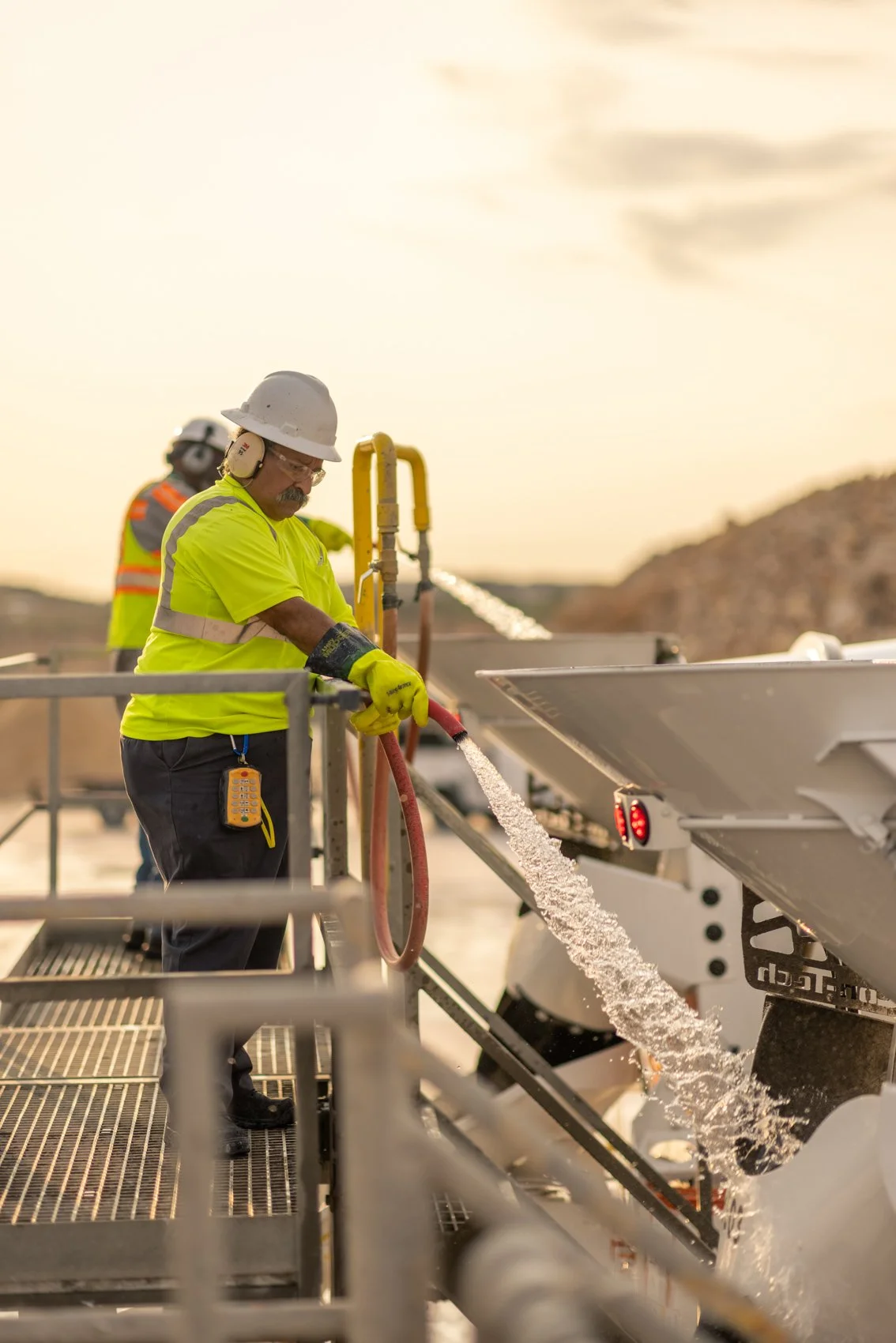 A worker in a yellow safety vest and hard hat stands on a platform, handling a hose that sprays water onto a large industrial machine, with another worker in the background and mountains in the distance during sunset.