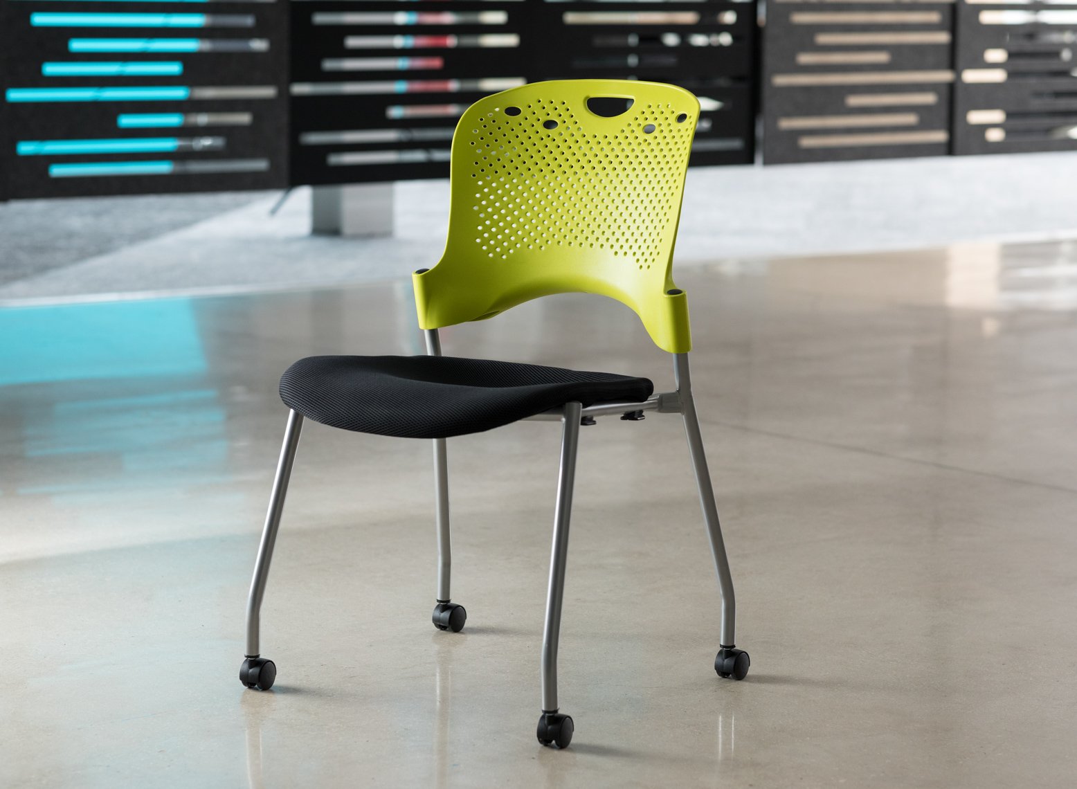 A modern office chair with a black seat, green backrest, and rolling caster wheels on a polished floor, with a dark background featuring a lit display.