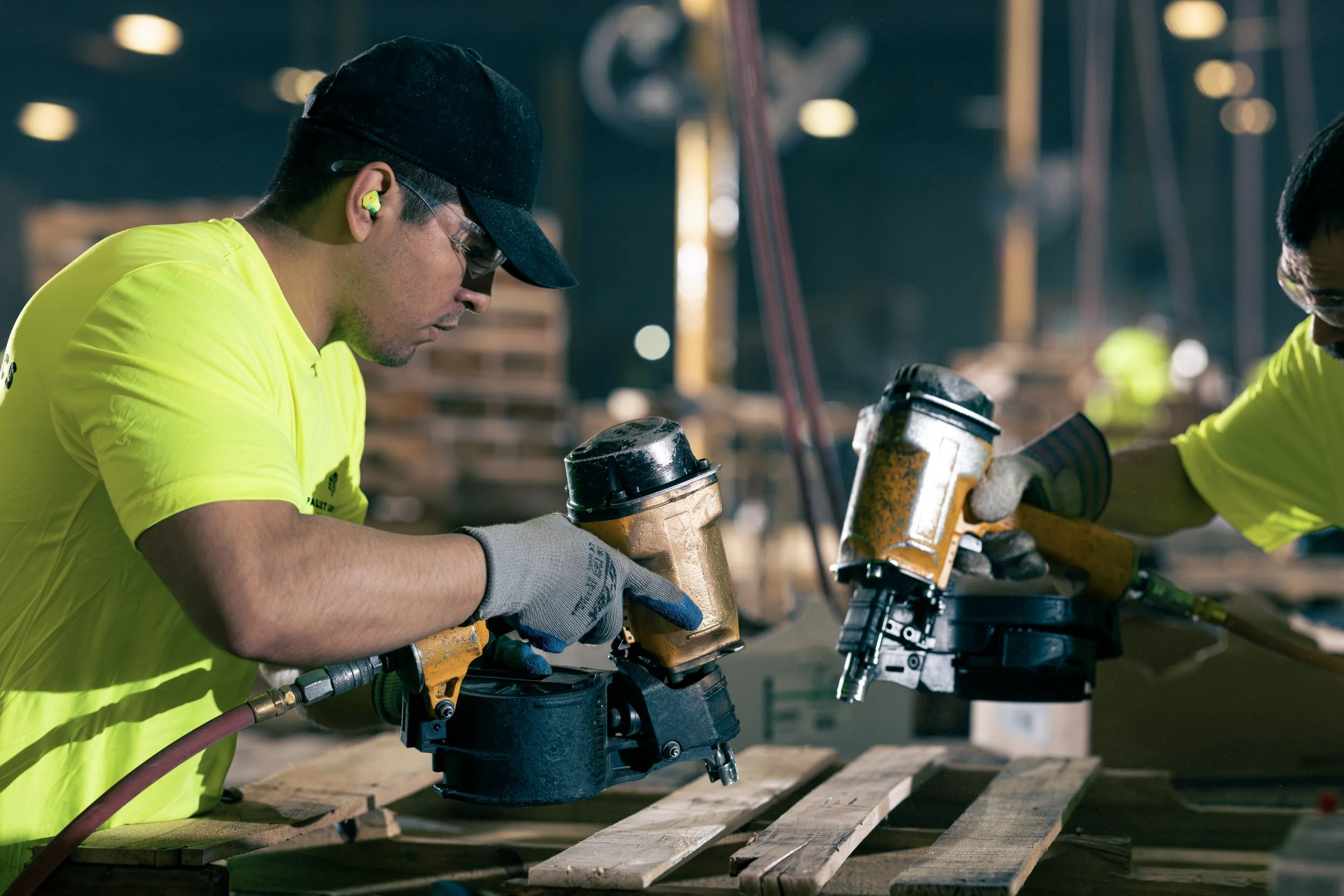 Worker in yellow shirt, wearing safety glasses and ear plugs, operating a handheld power saw on a wooden plank at a construction site.