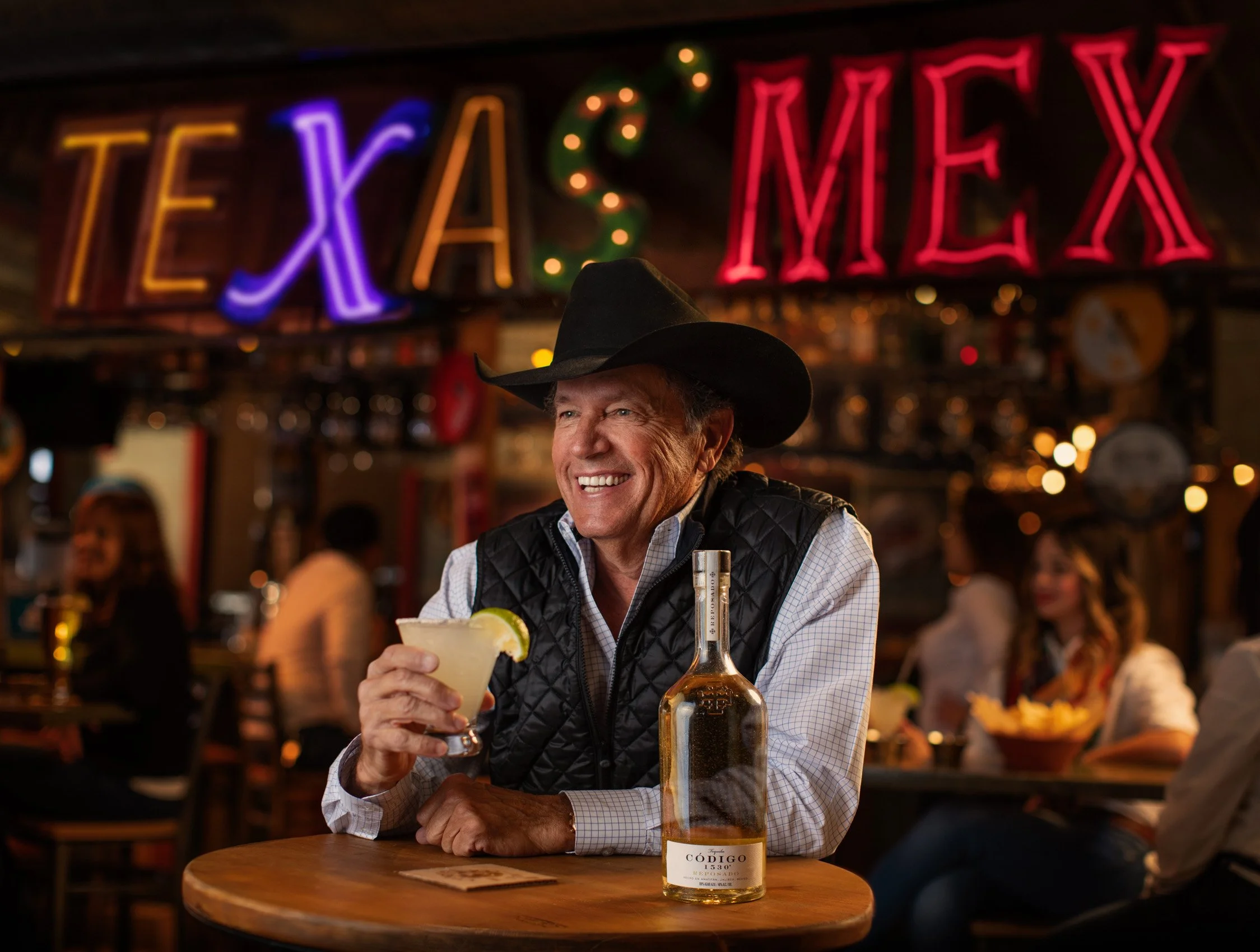 A smiling man wearing a cowboy hat, plaid shirt, and vest sitting at a wooden table in a bar or restaurant, holding a margarita glass with a lime wedge. There are colorful neon signs behind him, including the word "TEXAS" and other objects at the bar