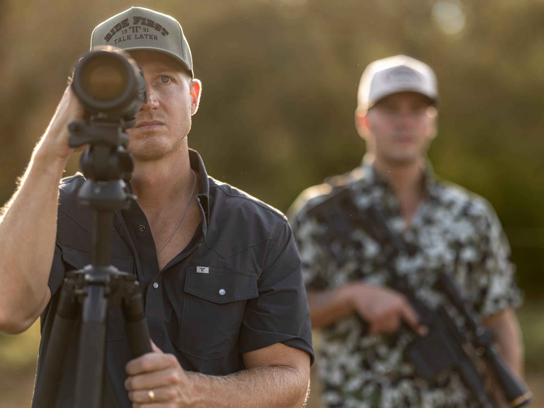 A man aiming a rifle at the camera with another person blurred in the background during sunset.