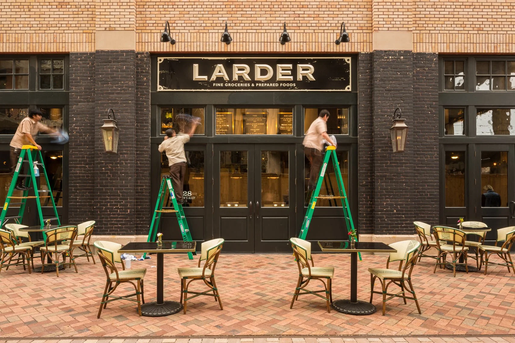 Workers on ladders cleaning the exterior of a restaurant named Larder with outdoor seating in front.