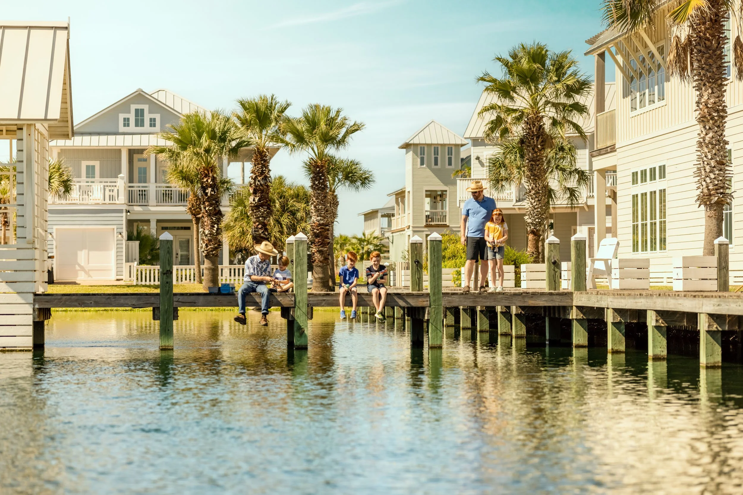 People sitting and walking on a wooden dock along a canal with houses and palm trees in the background, under a clear blue sky.