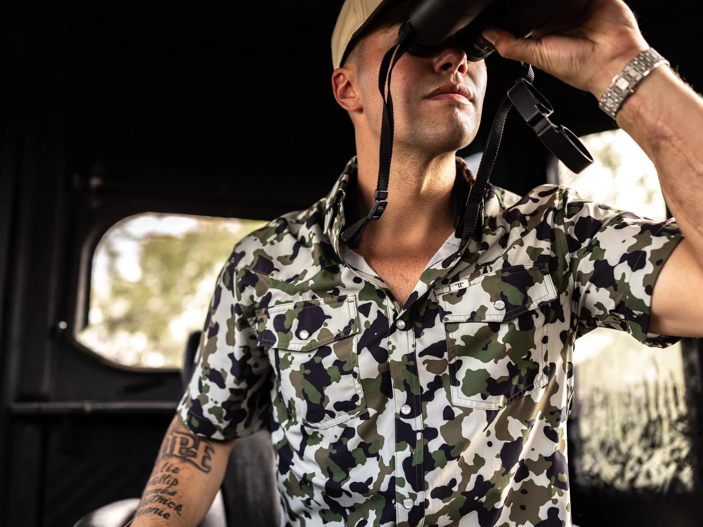 Man wearing camouflage shirt and watch, looking through binoculars, outdoors with blurred trees in background.