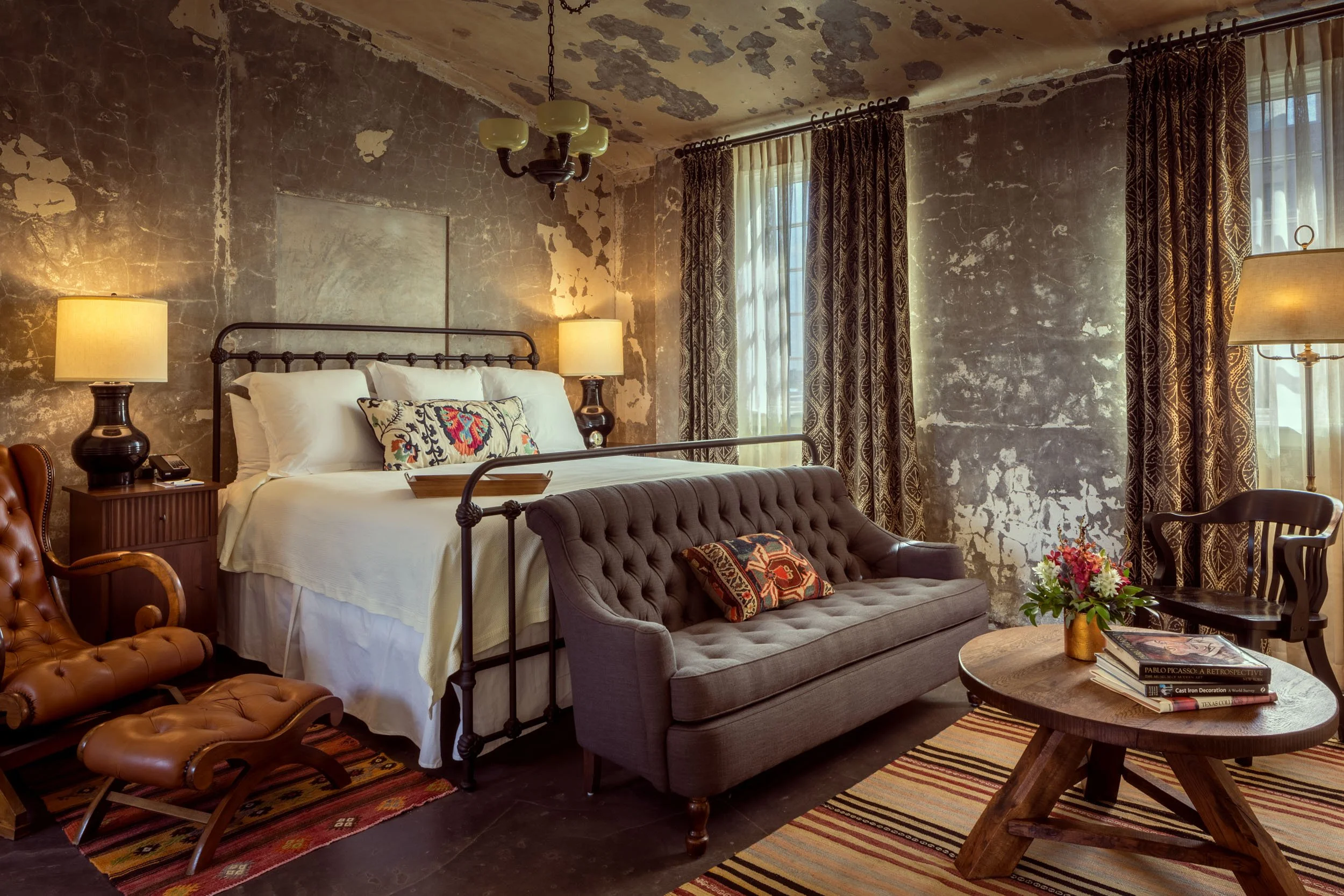 A rustic bedroom with a distressed, peeling wall finish. It features a bed with white linens and a decorative pillow, bedside tables with lamps, and a mix of vintage and modern furniture including a tufted leather armchair and a wooden coffee table w