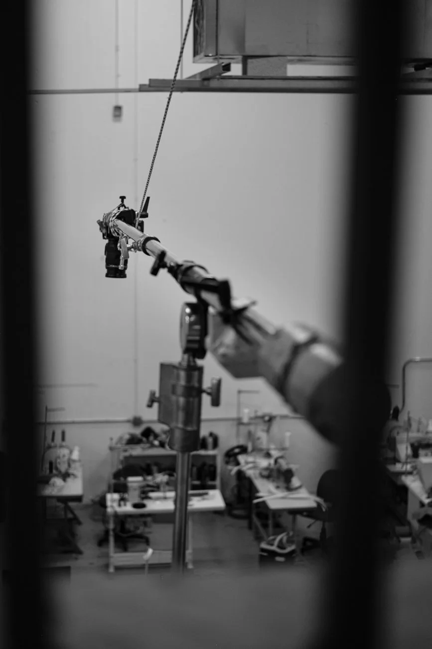 A black and white photo of a robotic arm in a laboratory or workshop, seen through a frame of vertical bars in the foreground, with tables and equipment in the background.