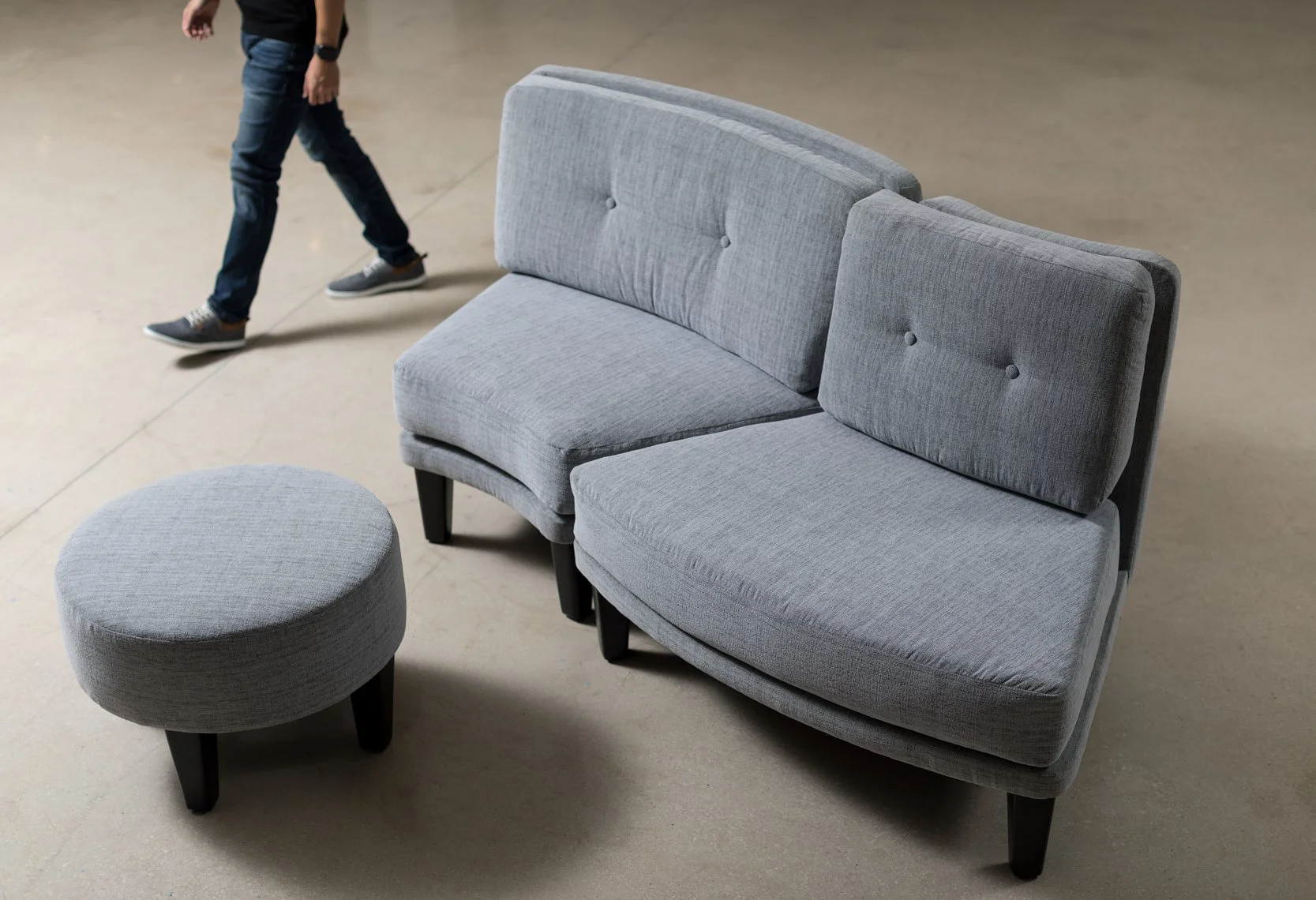 A light gray upholstered sectional sofa with black wooden legs, including a matching round ottoman nearby, situated on a smooth, beige floor in an indoor setting, with a person wearing jeans, a black shirt, and gray shoes walking past in the backgrou