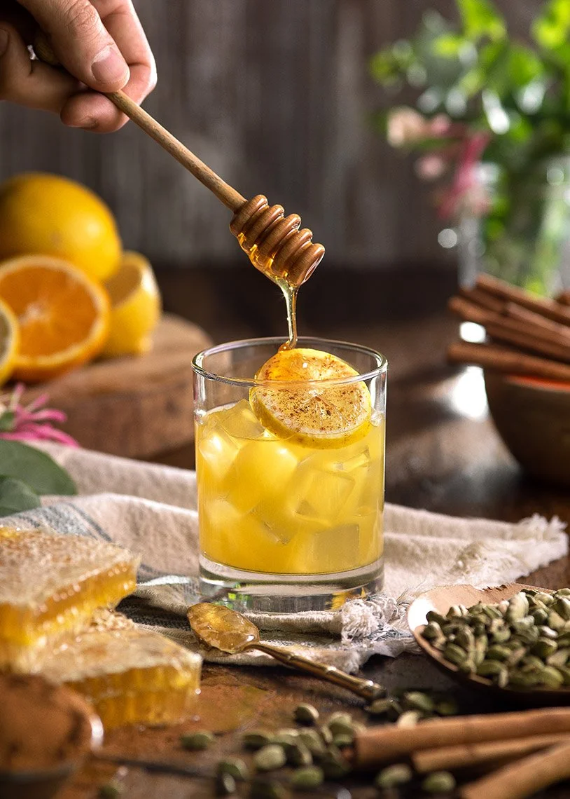 A glass of iced lemon honey tea with a lemon slice and honey dipper hovering above, honey dripping into the glass, surrounded by lemons, honeycomb, and spices on a rustic table.