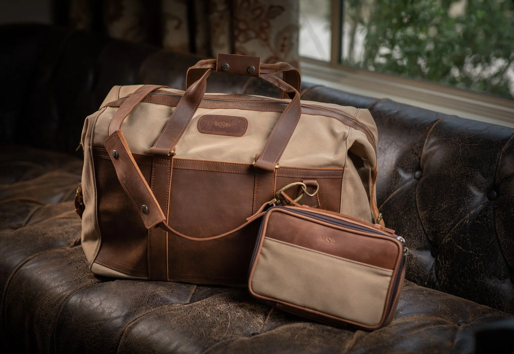 A beige and brown leather travel bag with handles, a detachable shoulder strap, and a front pocket, placed on a vintage leather sofa with a smaller matching pouch next to it. A window with greenery outside is in the background.