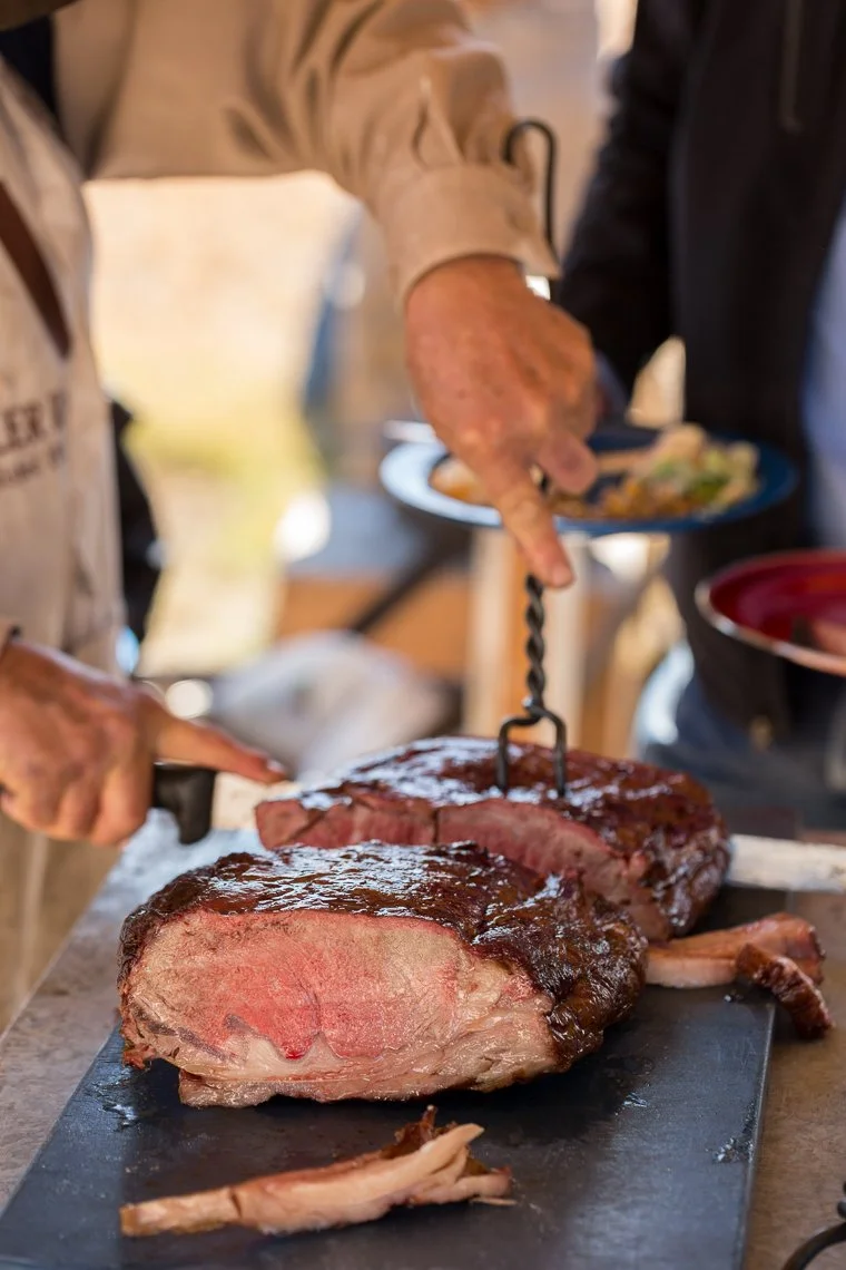Close-up of a cooked prime rib roast on a black cutting board, with a person carving it with a meat fork, in a casual dining setting.
