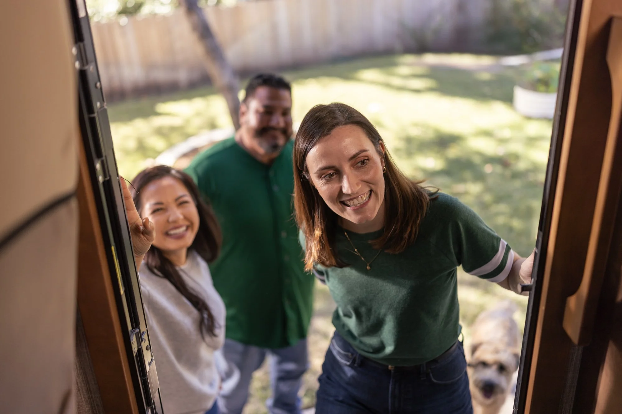 Three people peering through an opening, looking inside, in a backyard with trees and a fence. One woman is smiling at the camera, and a man behind her has a beard and is smiling. The third person is a woman with long hair, smiling.
