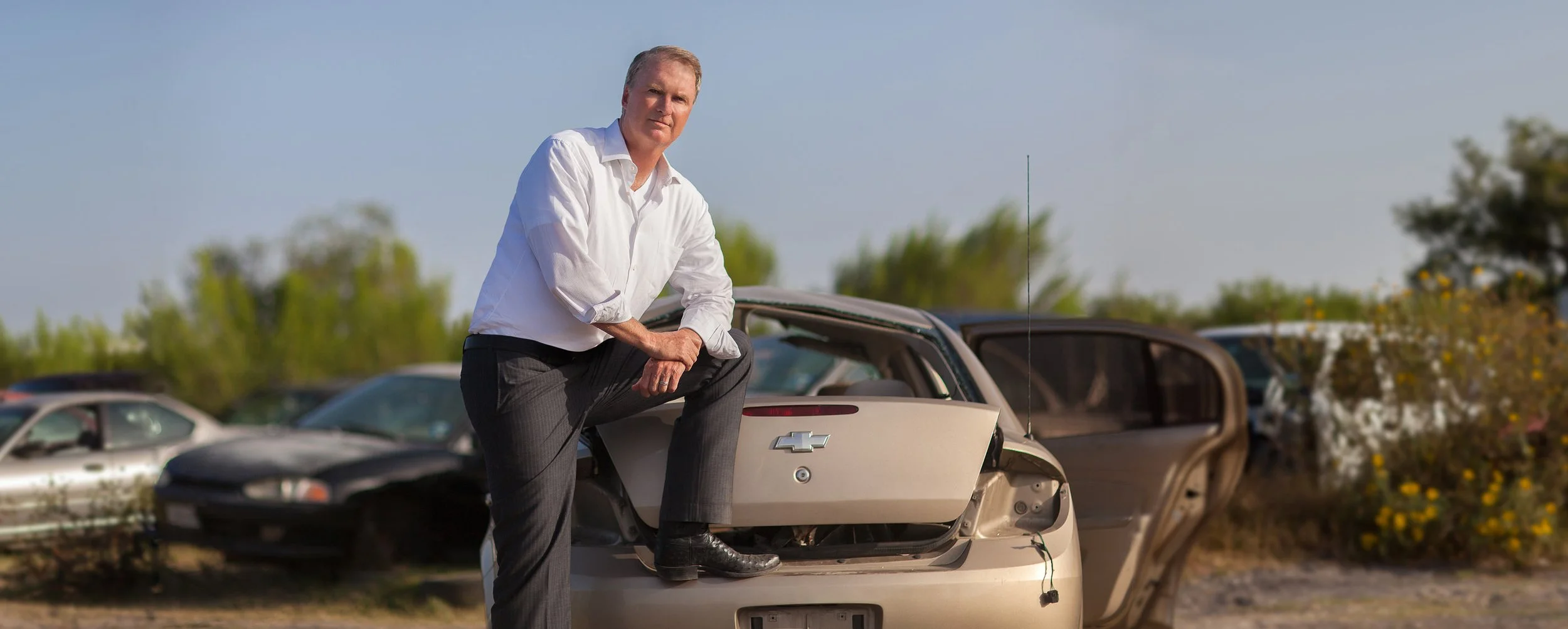A middle-aged man in a white dress shirt and dark pants posing on a damaged white Chevrolet car in a junkyard with other abandoned cars and trees in the background.