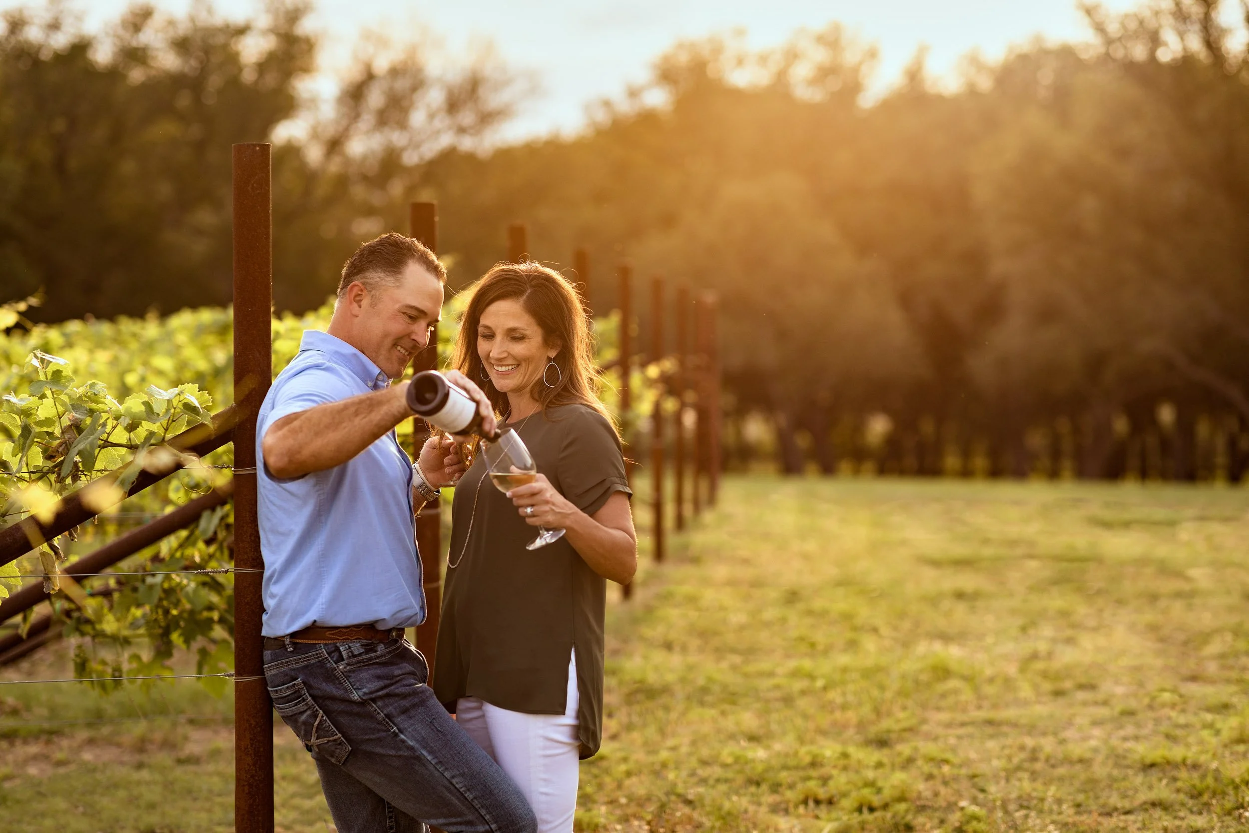 A man and a woman are standing in a vineyard, smiling and looking at a wine bottle. The man is pouring wine into a glass held by the woman, with sunlight shining through the trees in the background.