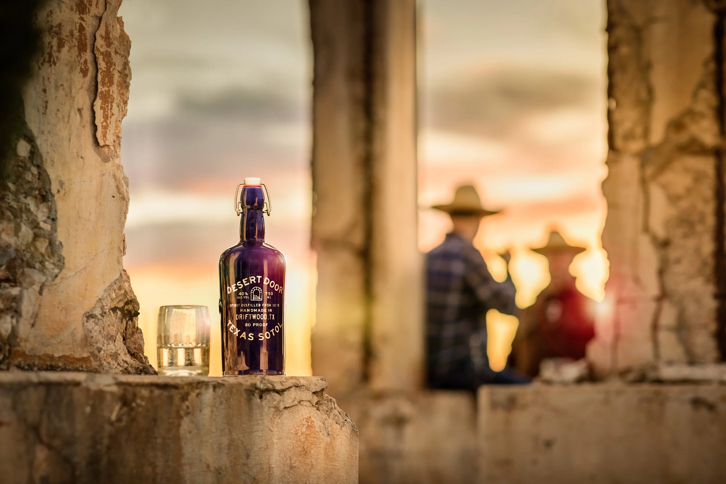 Close-up of a bottle of Desert Door Texas Sotol on a ledge with a glass in front of it. The background shows a blurred sunset and stone columns in an outdoor setting.