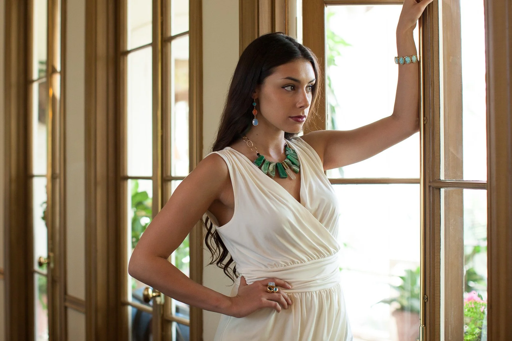A woman with long dark hair wearing a cream-colored dress and colorful jewelry stands by a wooden window, looking thoughtfully outside.