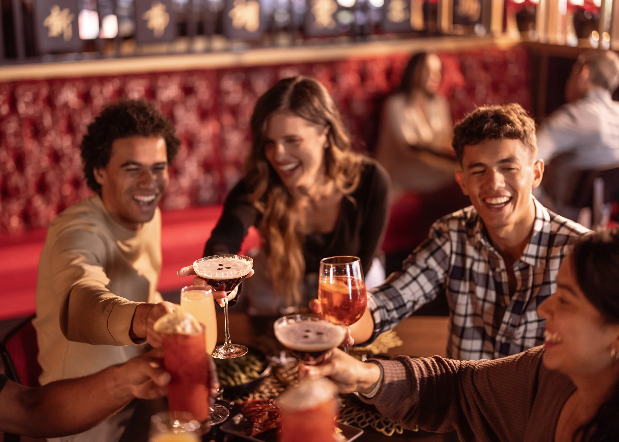 Group of friends sitting at a bar, smiling and toasting with various cocktails and drinks.