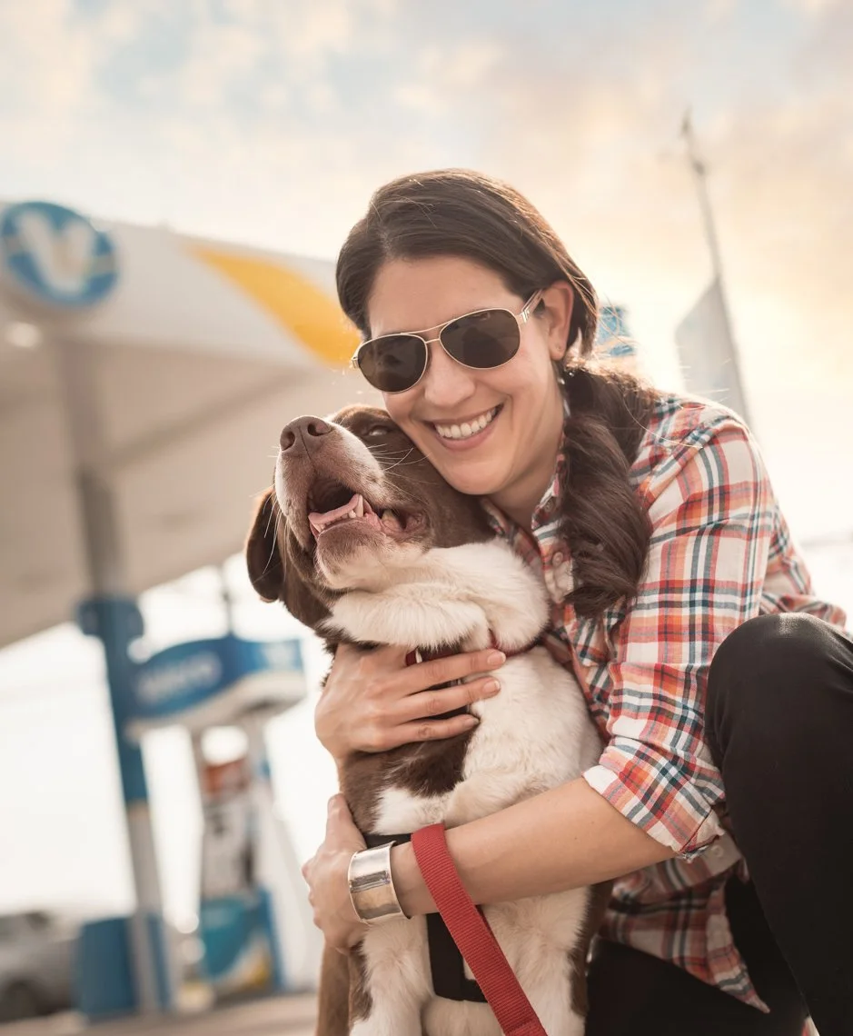 Woman smiling and hugging a puppy at a gas station, wearing sunglasses and a plaid shirt.