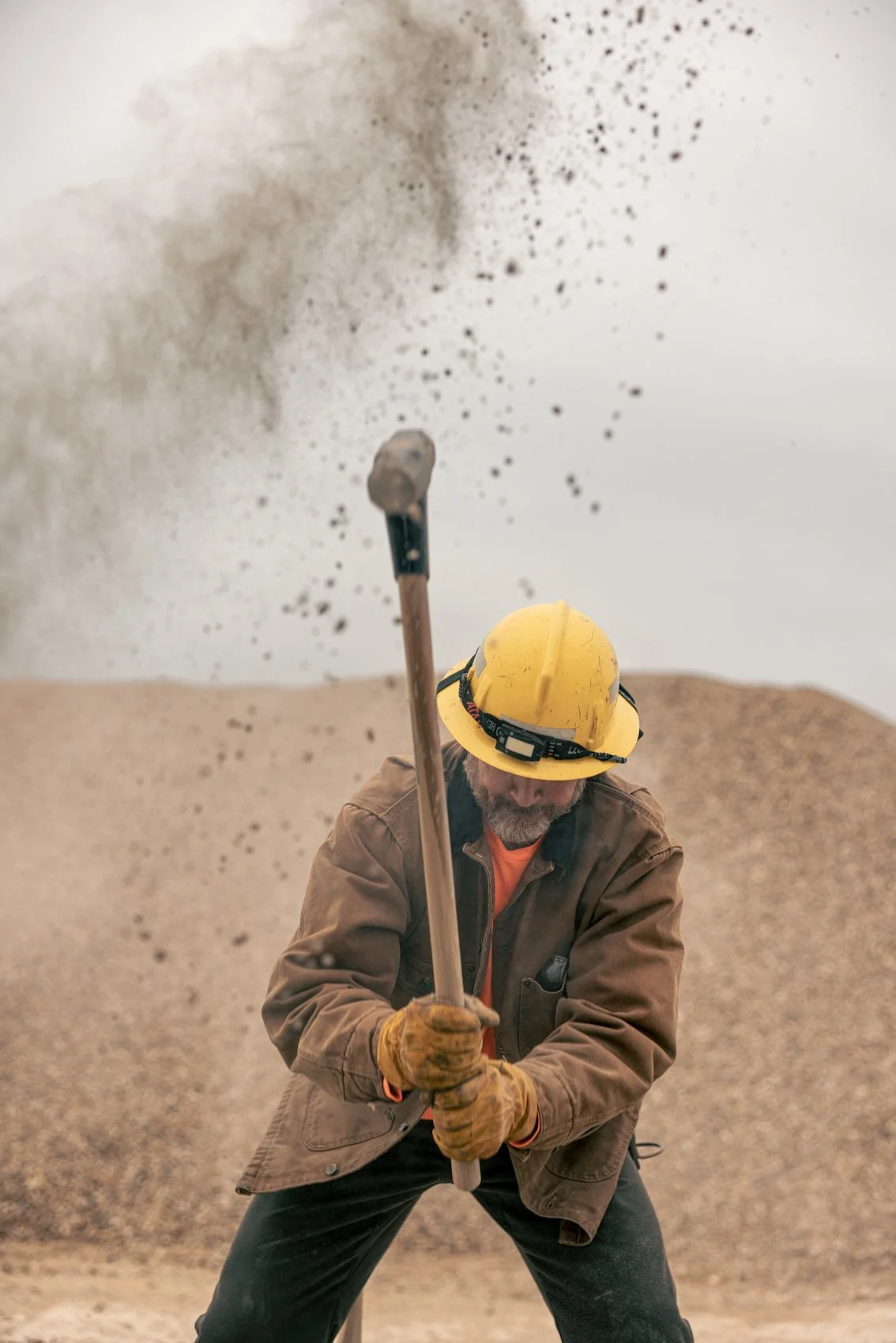 A construction worker with a yellow hard hat and brown jacket using a sledgehammer to break rocks, with dirt flying in the air in a quarry or excavation site.