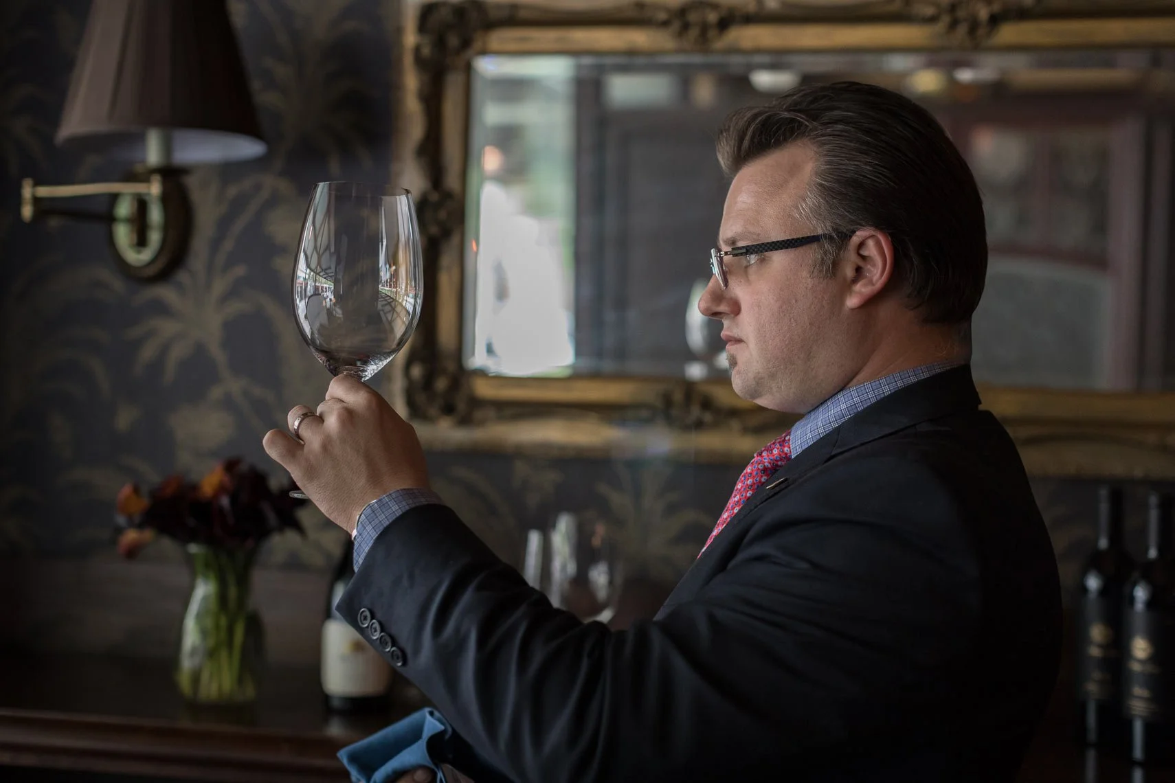 A man in formal attire, wearing glasses, holding a glass of red wine and examining it carefully.