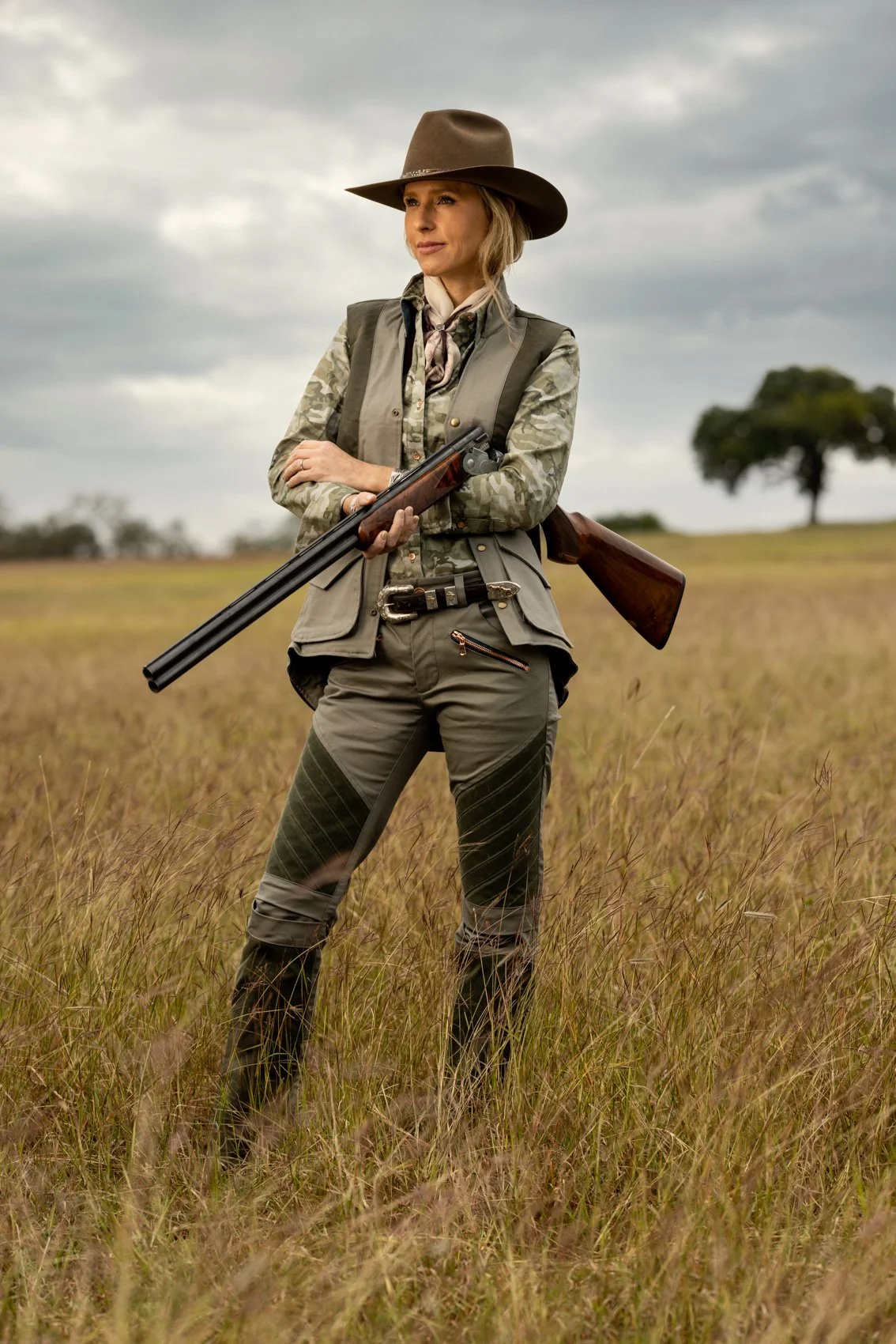 Woman dressed as a hunter standing in a grassy field with cloudy skies, holding a shotgun and wearing a wide-brimmed hat.