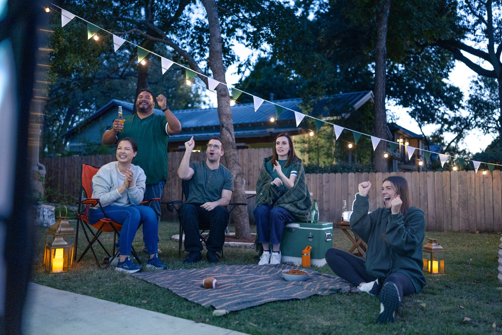 A group of friends enjoying an outdoor evening gathering, sitting on camping chairs and a blanket in a backyard, decorated with string lights and lanterns. Some people are cheering and holding drinks, with trees and a house in the background.
