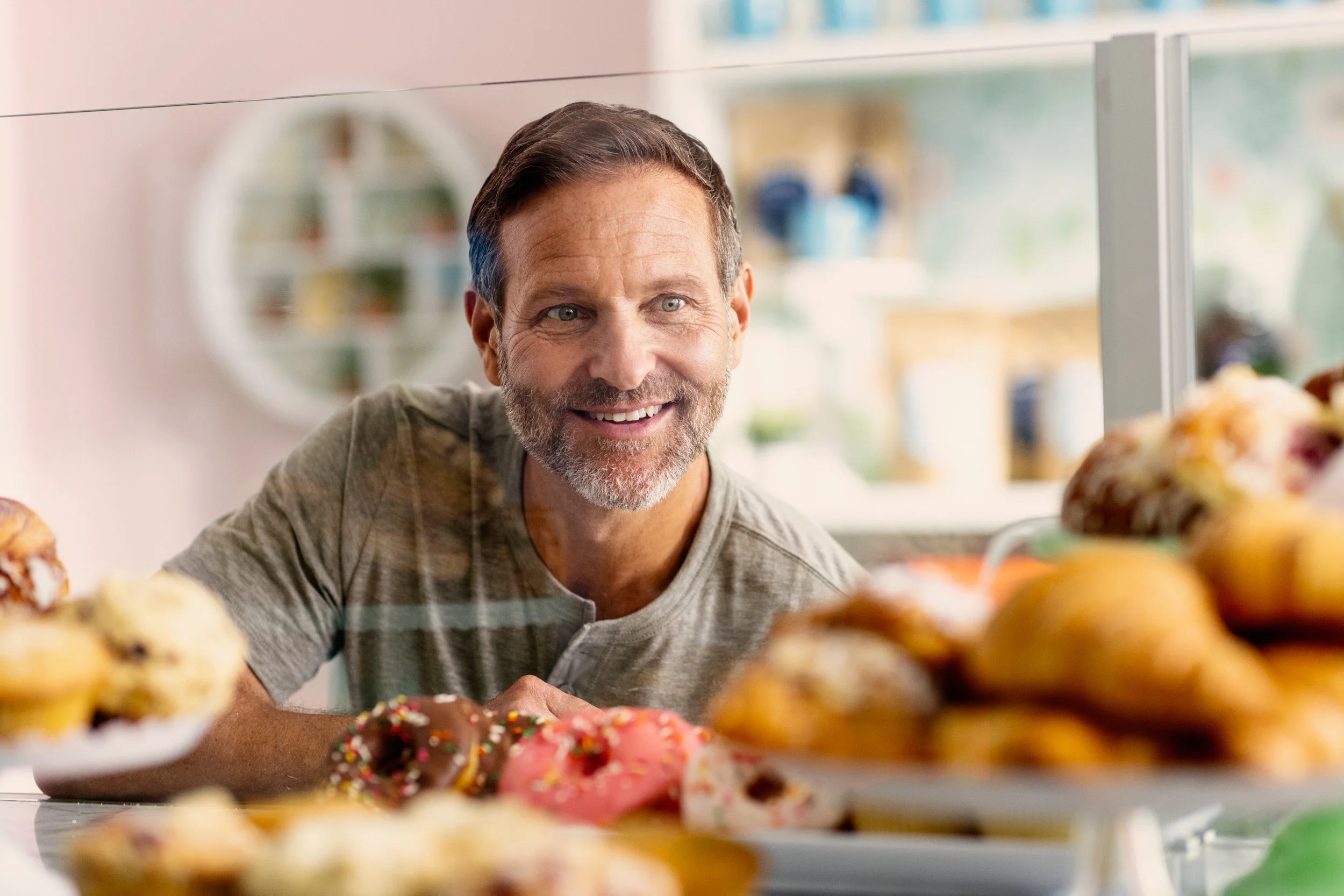 A man with gray hair and a beard smiling at a display of colorful donuts with sprinkles and frosting, in a bakery or kitchen setting.