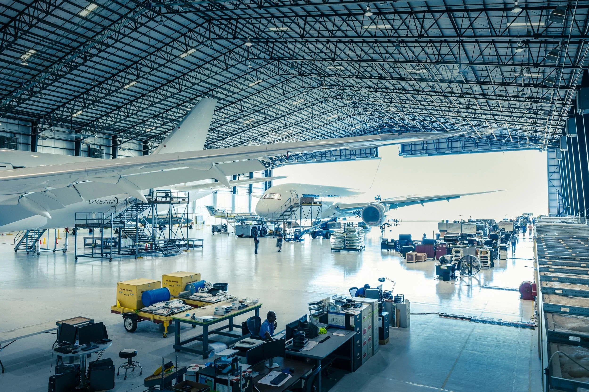 Inside an airplane hangar with large aircraft, equipment, carts, and workers preparing for flight or maintenance.