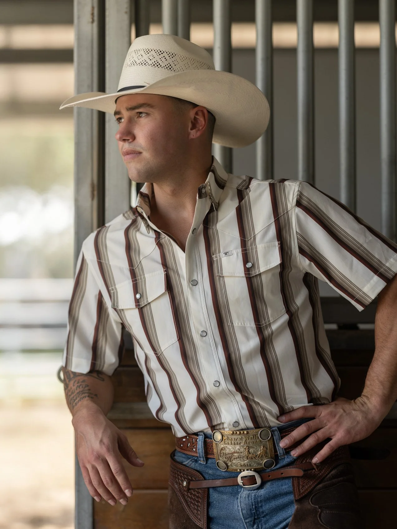 Man wearing a plaid western shirt, cowboy belt with large buckle, and cowboy hat, leaning against a wooden fence.