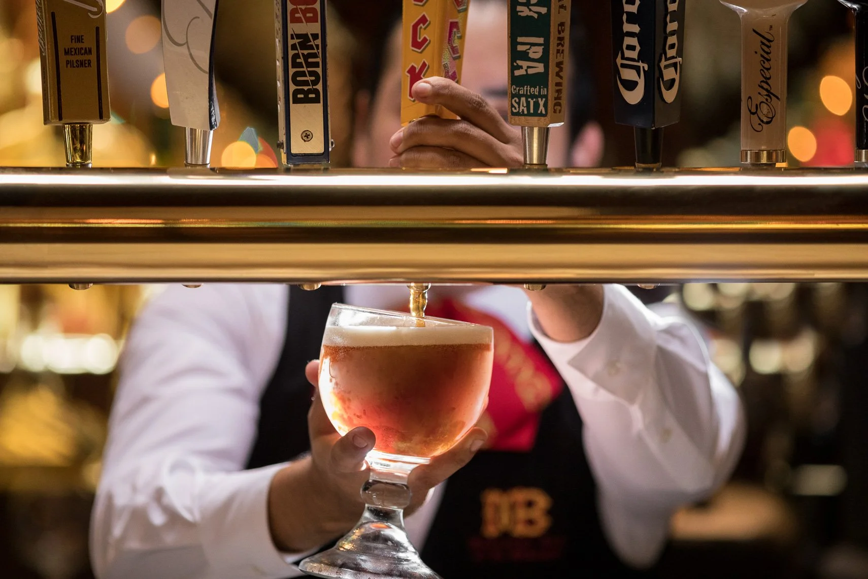 A bartender pouring a beer from taps into a glass, with a person holding the glass in the foreground. The background is blurred with warm lighting.