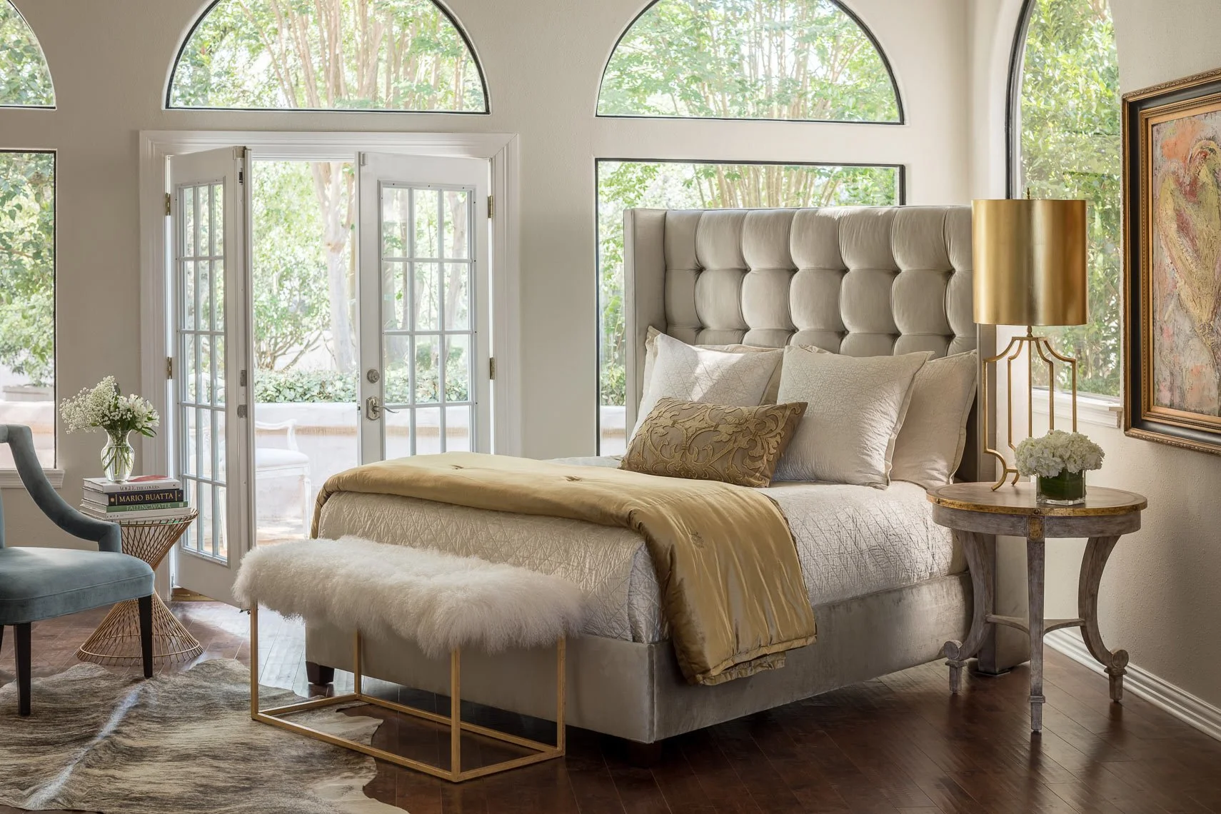 A bedroom with large arched windows and French doors leading to a patio, a tall beige upholstered headboard, a gold table lamp, a small round bedside table with flowers, and a bed with pillows and a blanket.
