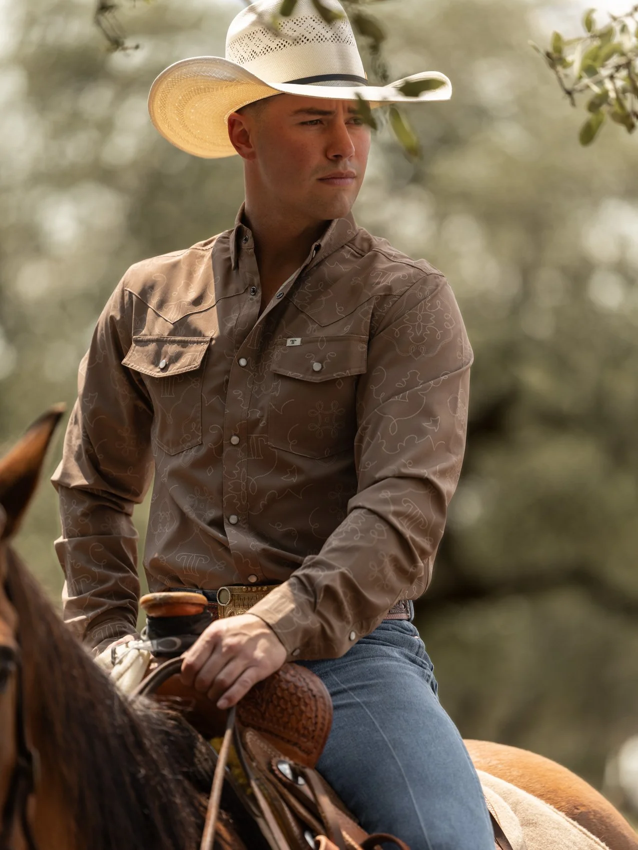 A young man wearing a cowboy hat, brown western shirt with embroidery, jeans, and a belt, riding a horse outdoors with blurred trees in the background.