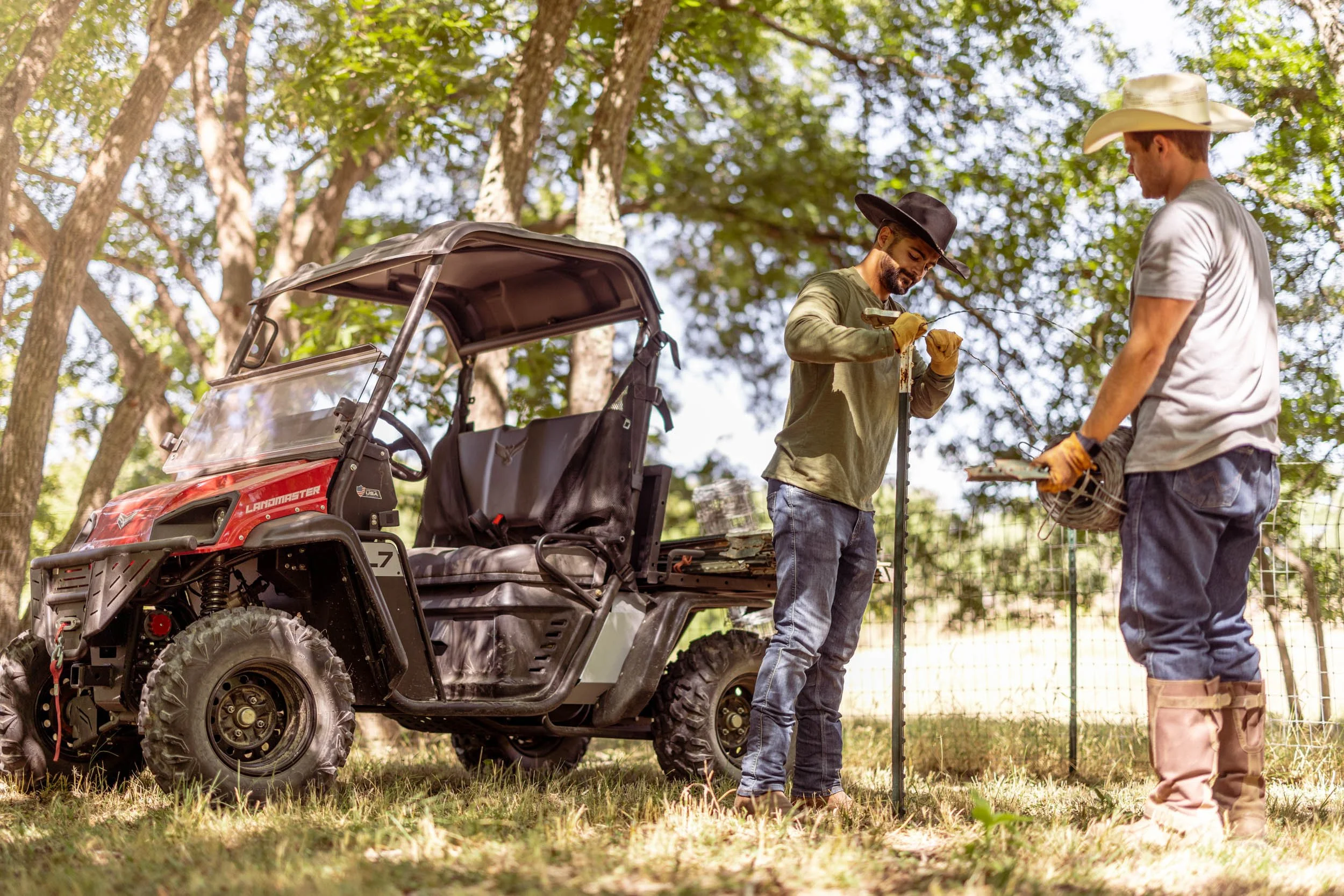 Two men wearing hats and gloves working together outdoors near a utility vehicle, with trees in the background.