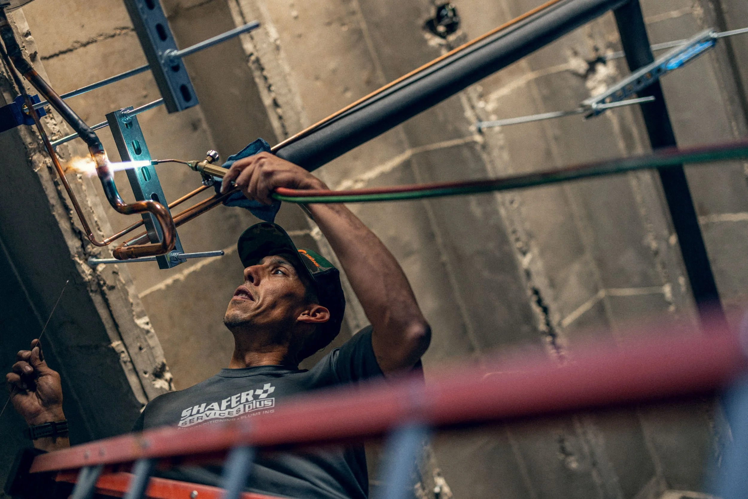 A man welding copper pipes against a concrete wall, wearing a safety cap and a dark shirt with white text.