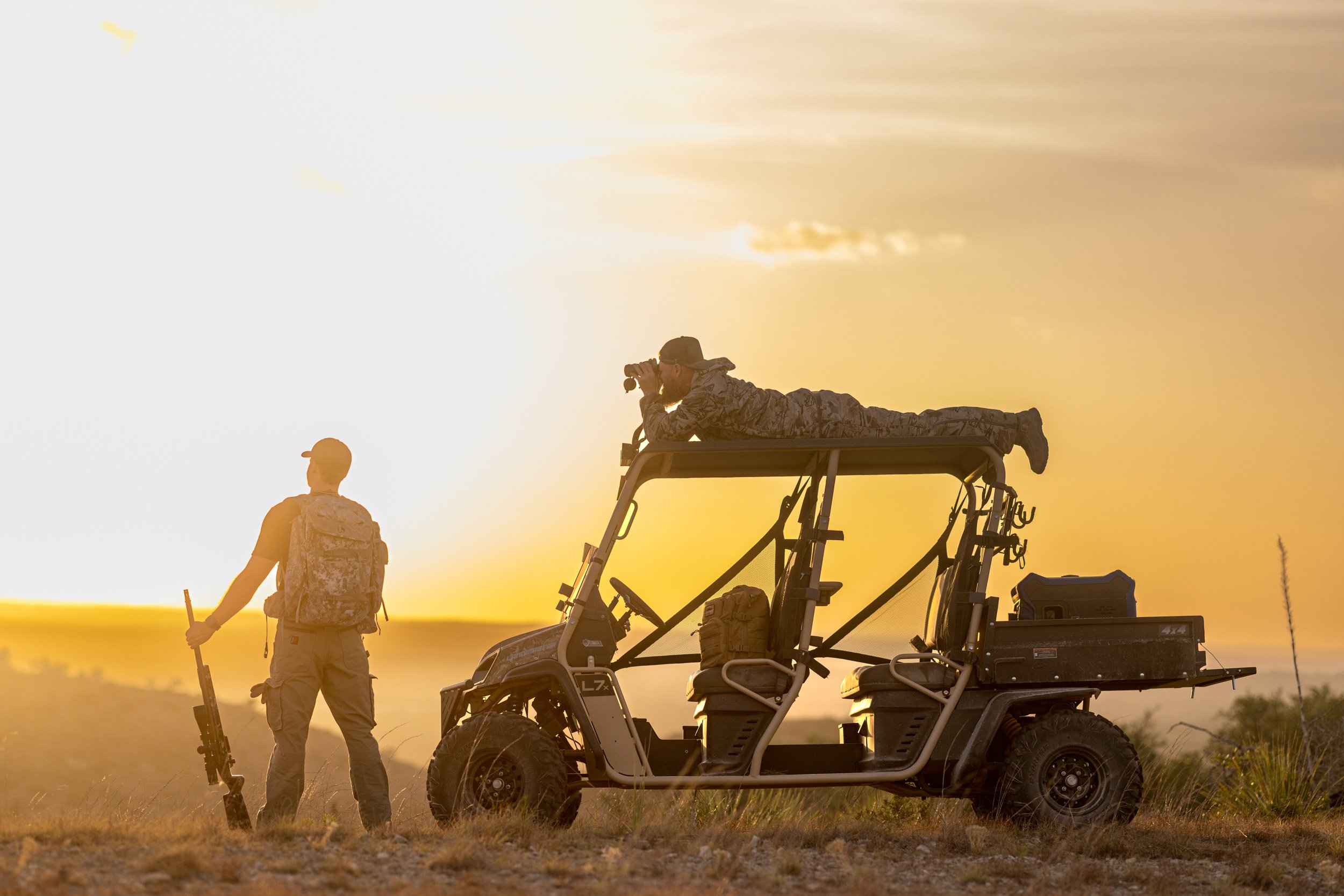 Two military personnel in camouflage uniform, one standing with a rifle, the other lying on a utility vehicle, during sunset with a vast open landscape.
