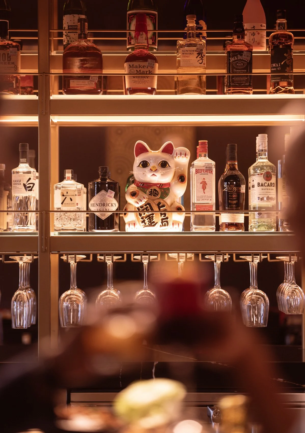 A bar shelf displaying various liquor bottles, a ceramic lucky cat figurine, and upside-down wine glasses hanging below the shelf.