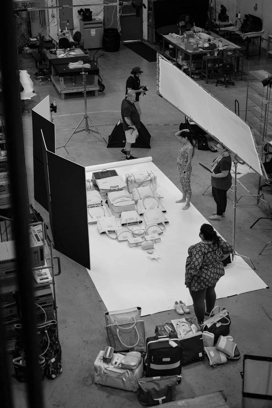 A behind-the-scenes view of a photo shoot setup with props laid out on a white backdrop, including various bags and accessories. Several people are preparing or organizing, with one person reading a script or notes, in an industrial-style studio with