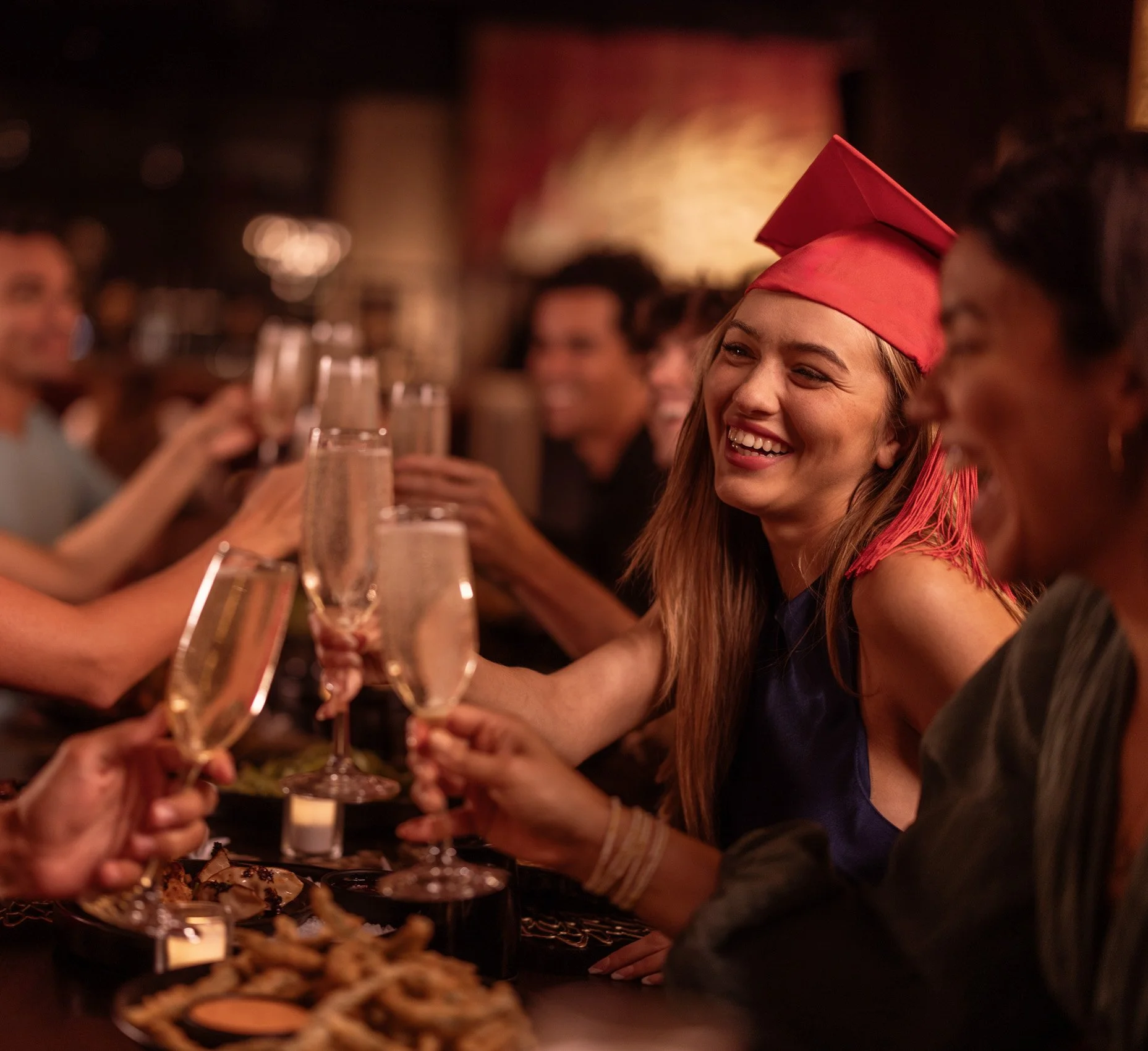 People celebrating at a party, with a woman wearing a red graduation cap and smiling, holding a champagne flute, with others around her also holding drinks, in a warmly lit setting.