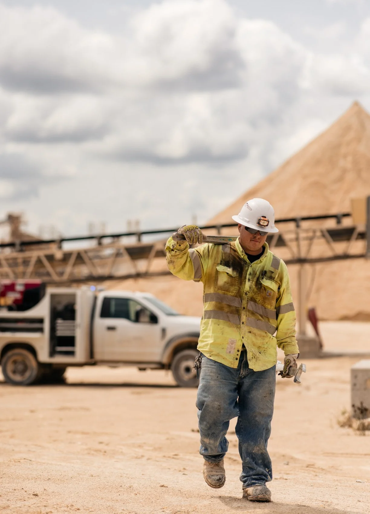 A construction worker wearing a yellow safety jacket, hard hat, sunglasses, and jeans walking across a construction site. He is carrying a tool over his shoulder, and there is a white pickup truck and a large mound of sand in the background under clo