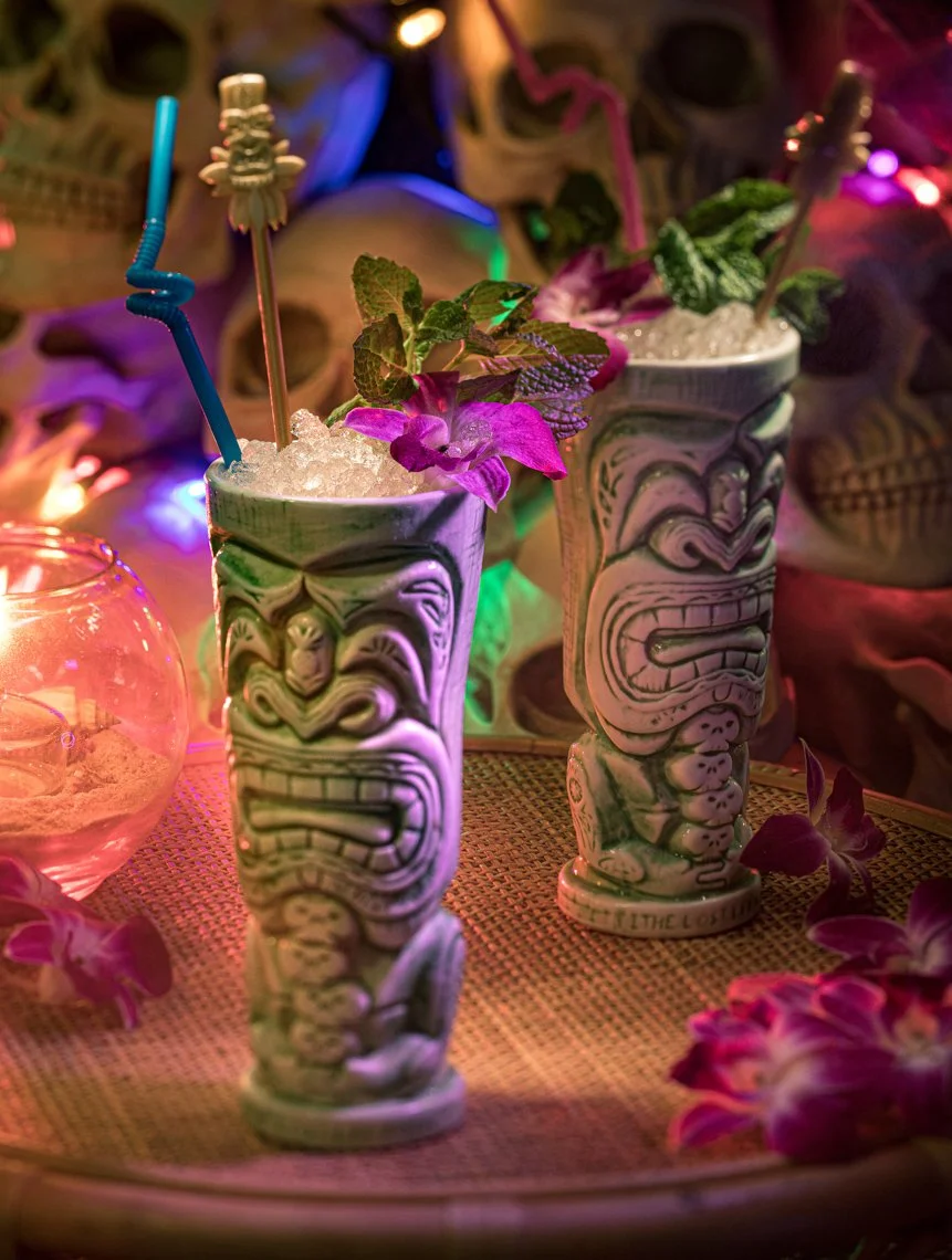 Two tiki-shaped ceramic cups filled with crushed ice and garnished with purple flowers and green leaves, with colorful lighting in the background.