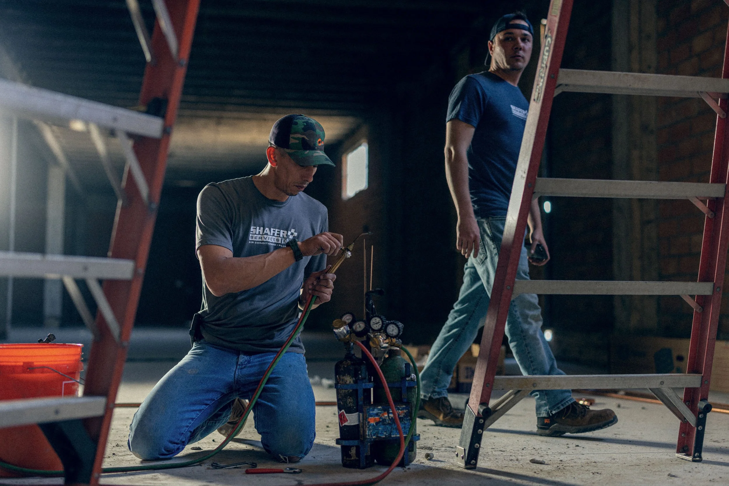 Two workers are installing or maintaining HVAC equipment inside a building. One worker is kneeling, holding tools, and wearing a camouflage cap and a shirt with the company logo 'Shafer Services.' The other worker is standing on a ladder, holding a t
