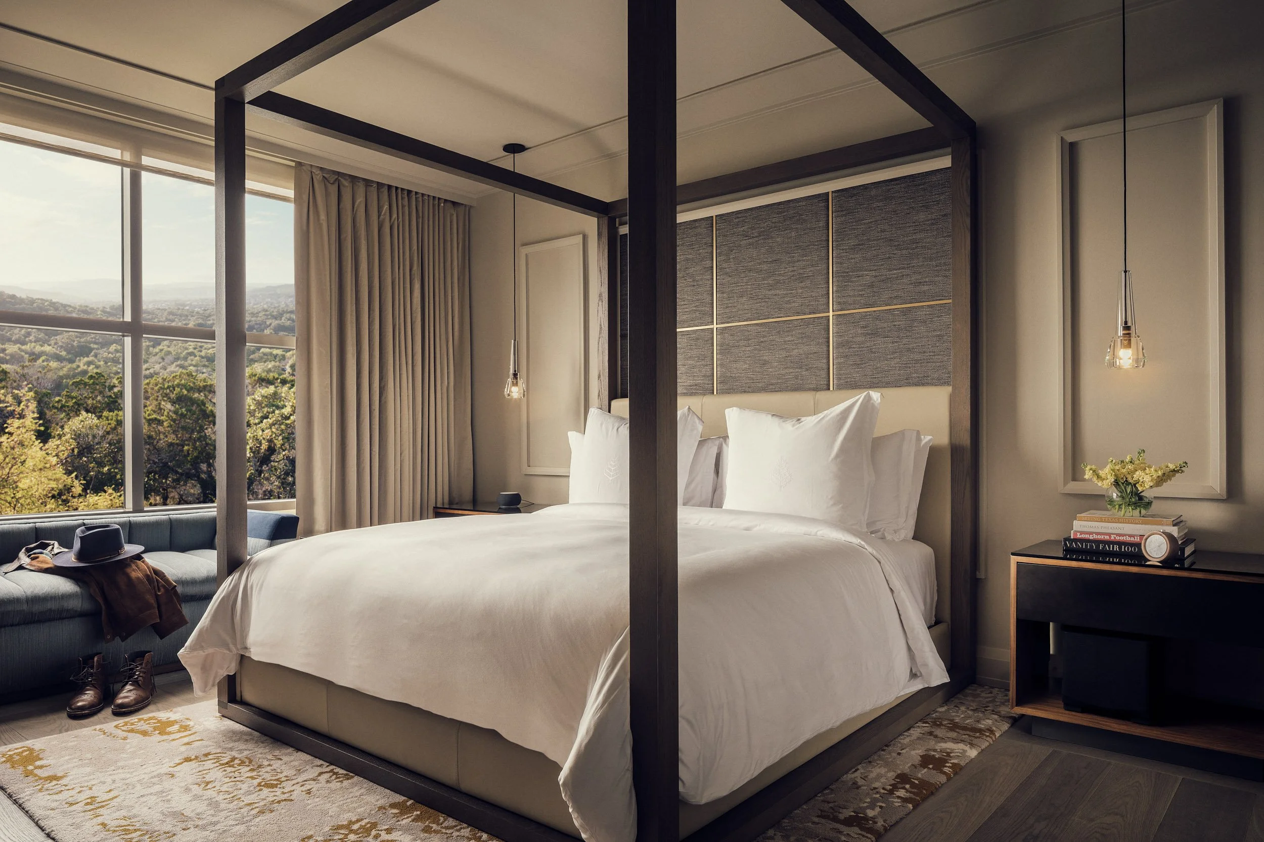 A modern hotel bedroom with a large bed and a view of trees and mountains through large windows, decorated with a hanging pendant light and books on a side table.