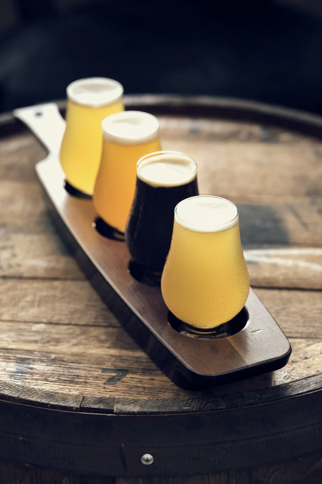 A wooden tray with four glasses of beer, two with pale yellow beer and two with dark stout, placed on a rustic wooden surface.
