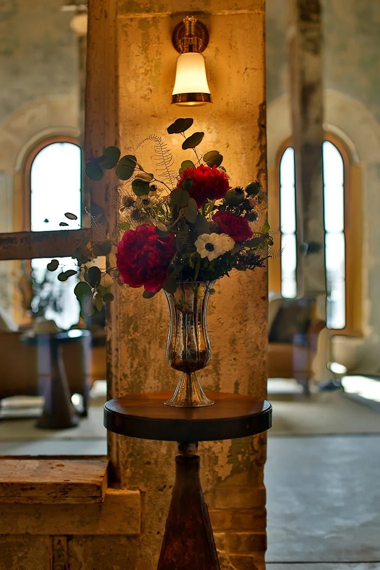 A tall glass vase filled with red, white, and green flowers on a round wooden table, set against an aged wall with a wall-mounted light fixture overhead, in a warmly lit interior space.