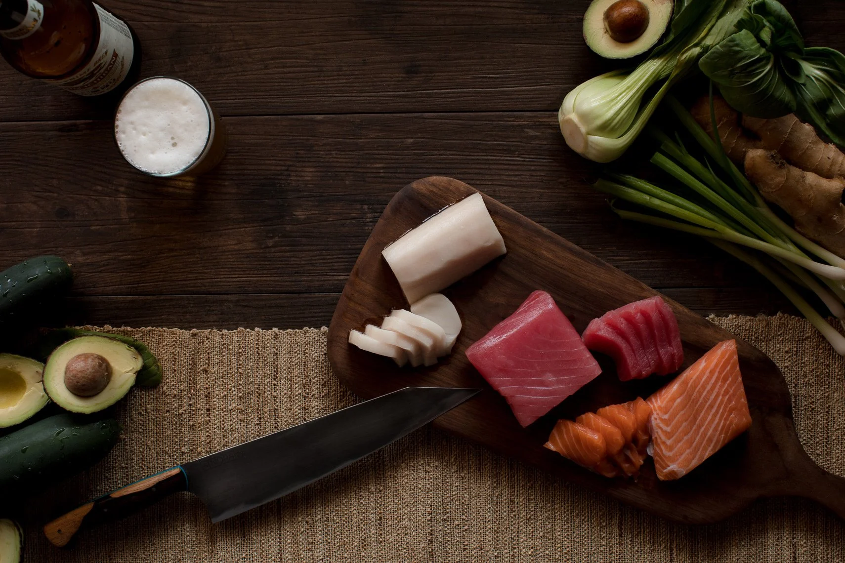 Sliced raw fish (salmon, tuna, and white fish) on a wooden cutting board surrounded by vegetables and other ingredients.