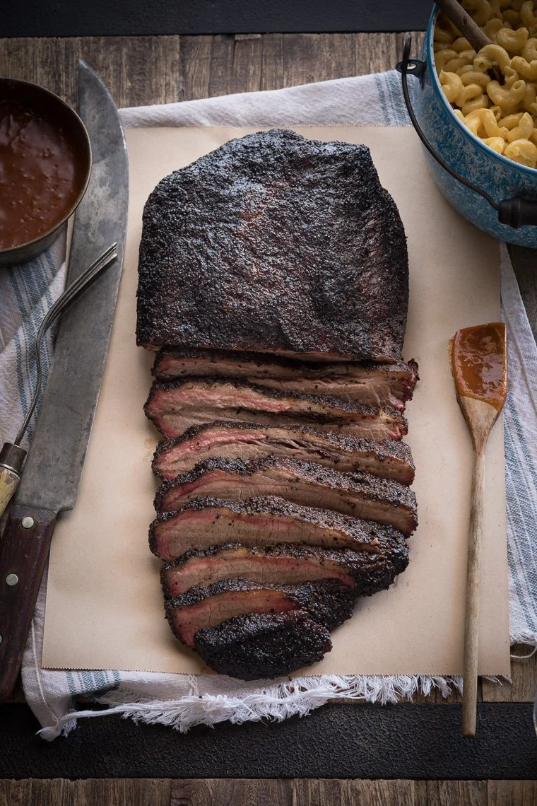 Sliced smoked brisket with a dark bark on a cutting board, served with barbecue sauce and macaroni and cheese in a bowl.